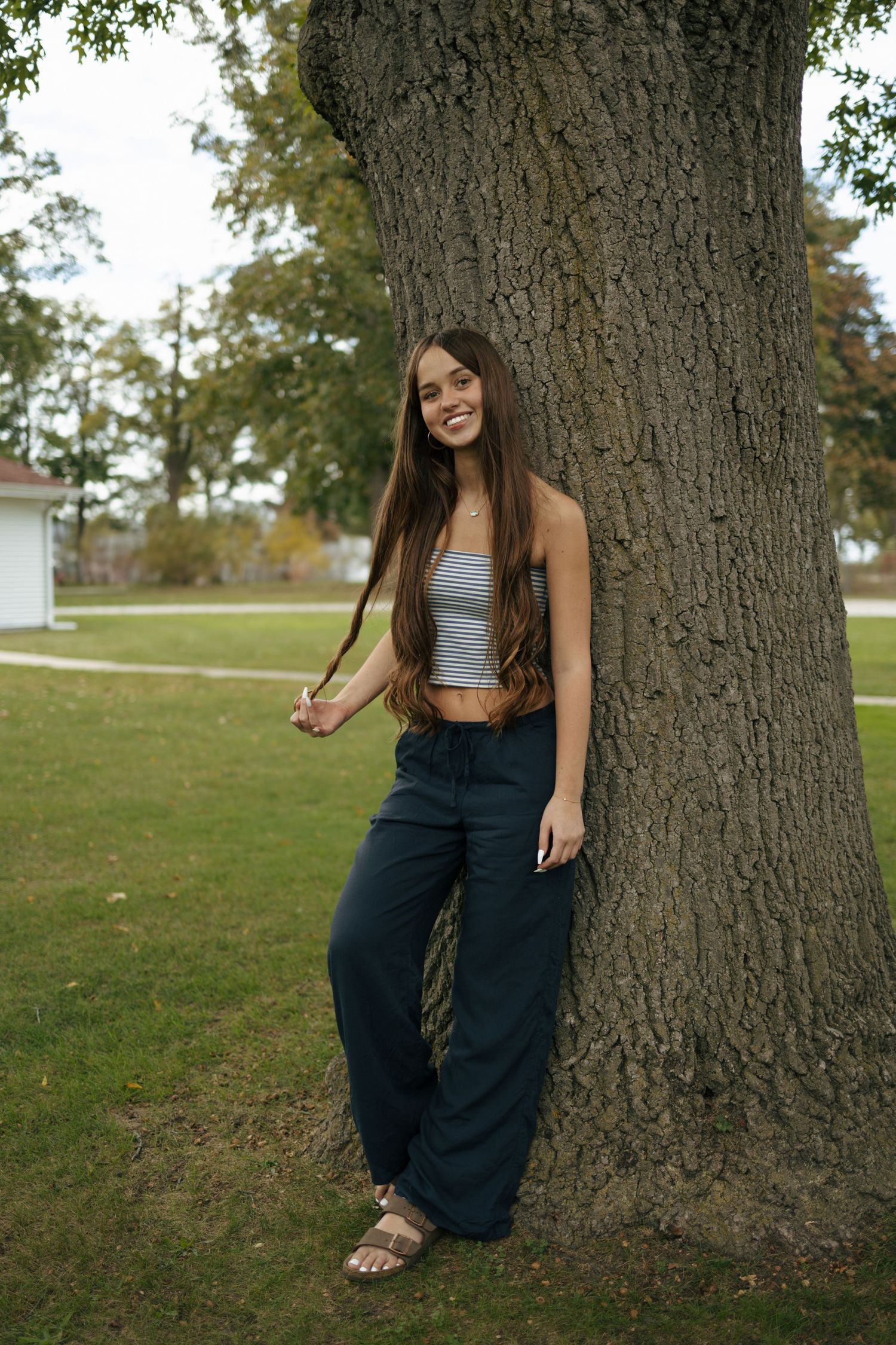 Young woman with long hair leaning against large tree trunk in park, wearing grey crop top and navy pants.