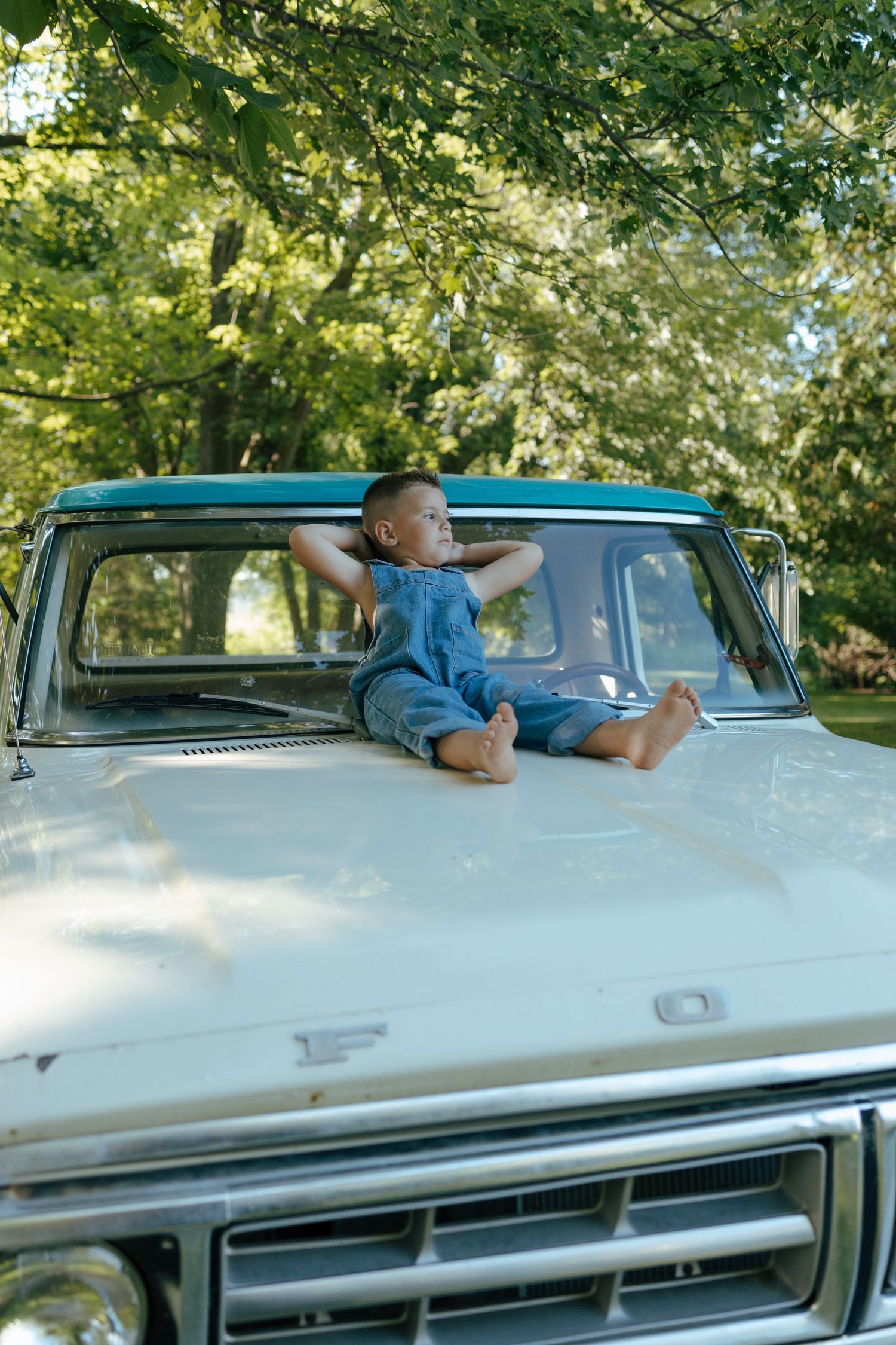 Barefoot boy in denim overalls relaxes on hood of vintage Ford truck under green trees.