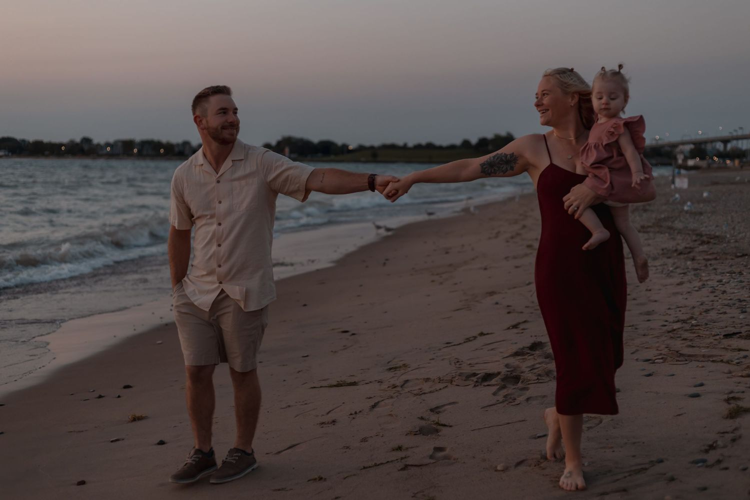 A smiling man reaches out to hold hands with a woman carrying their baby on a beach at sunset.