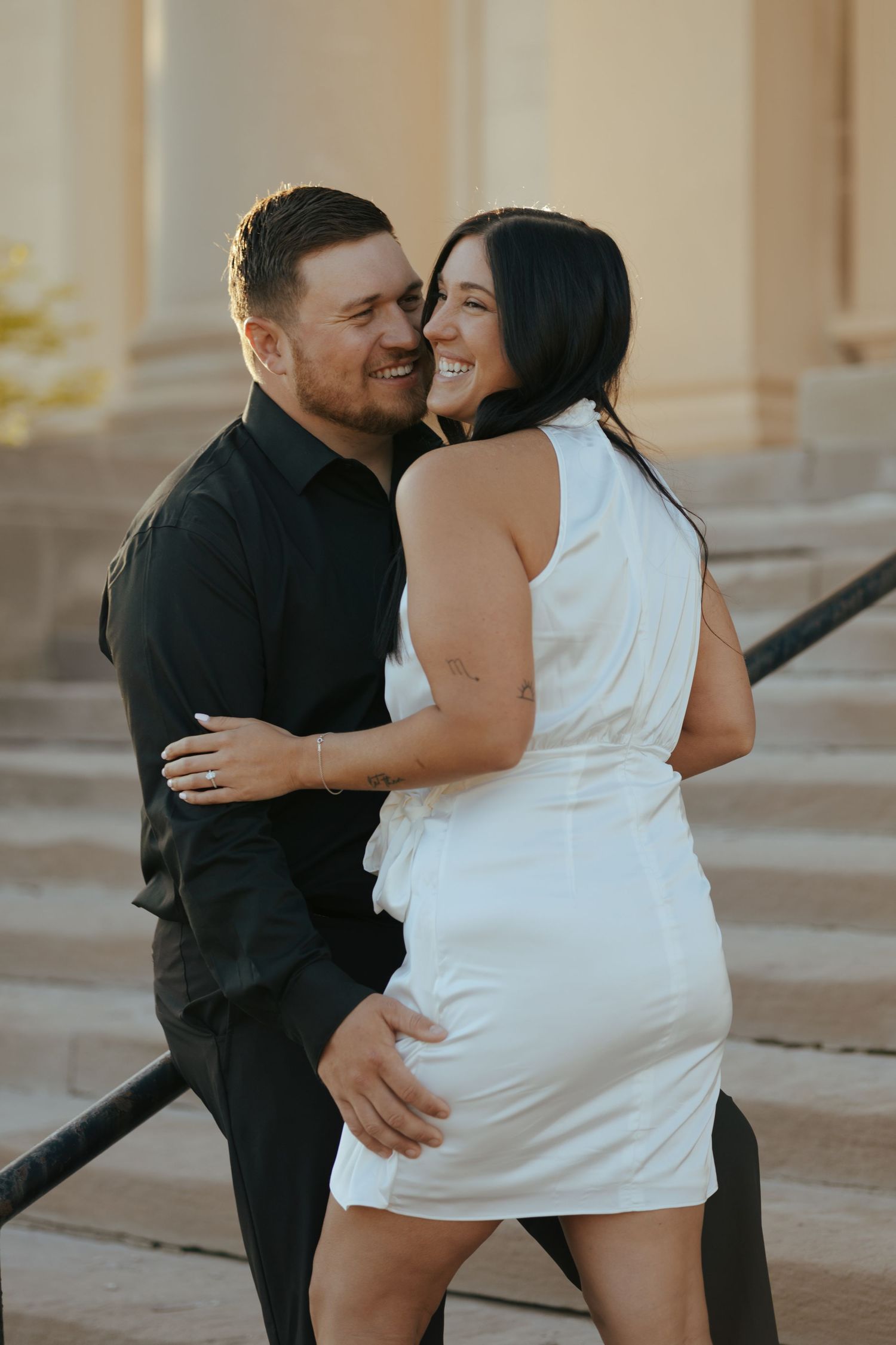 Laughing couple embracing on outdoor steps at golden hour in black and white outfits.