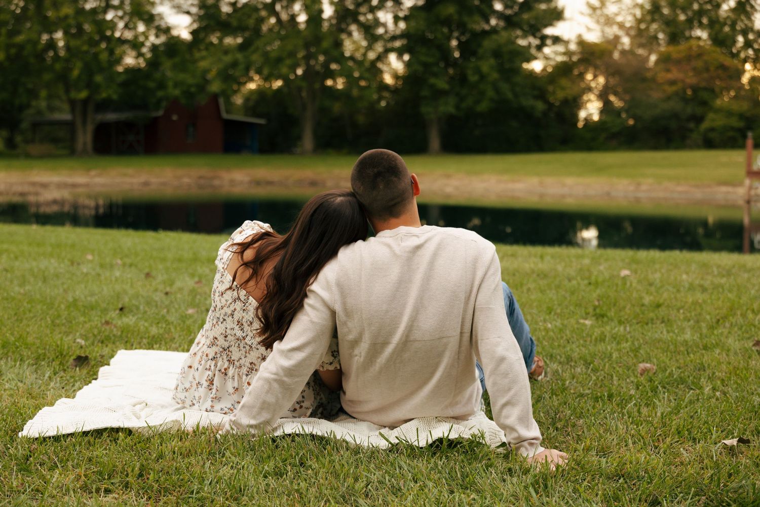 Couple sitting together on a blanket facing a serene pond in a green park at sunset.