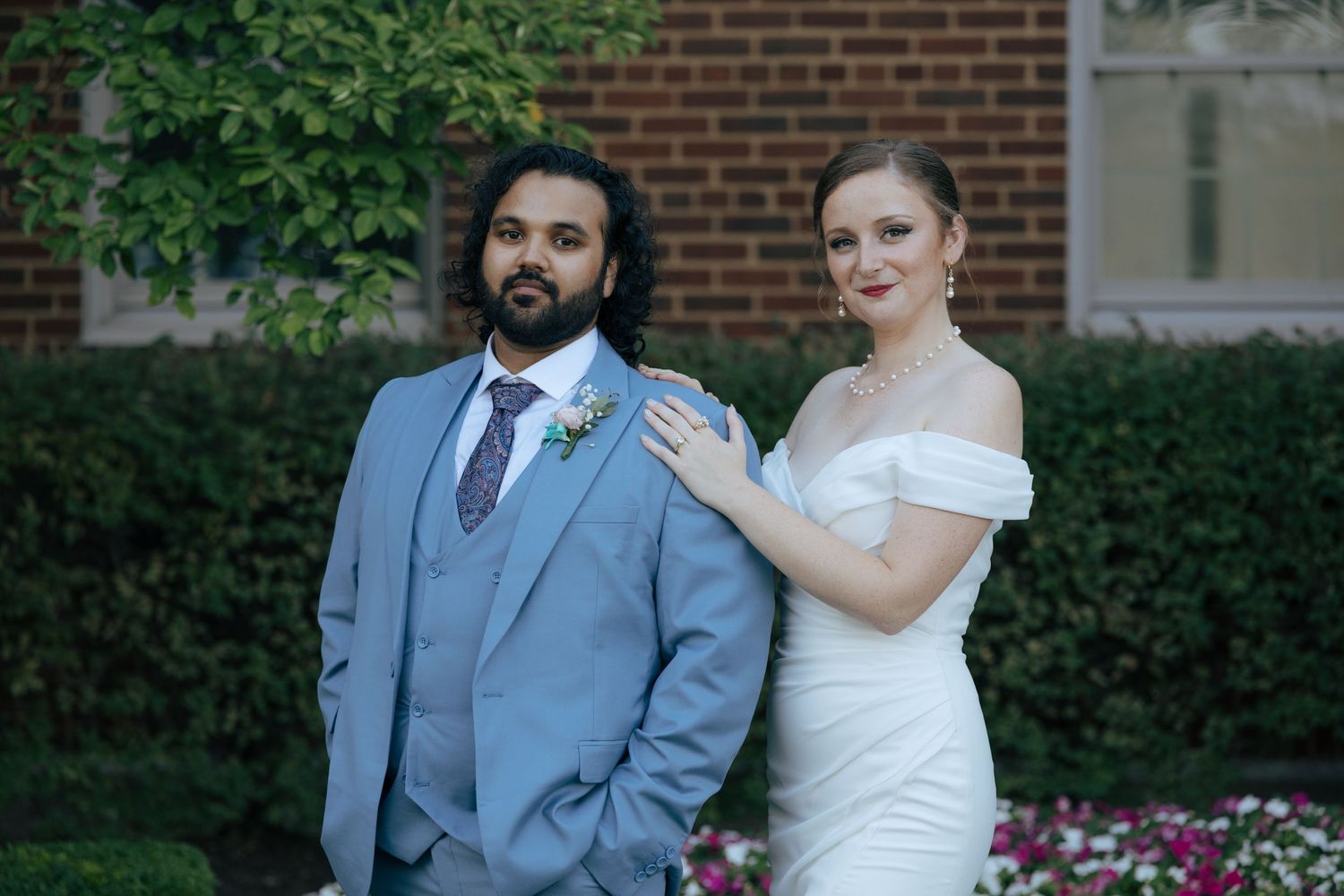 Bride in off-shoulder white gown and groom in light blue suit posing outdoors near brick building.