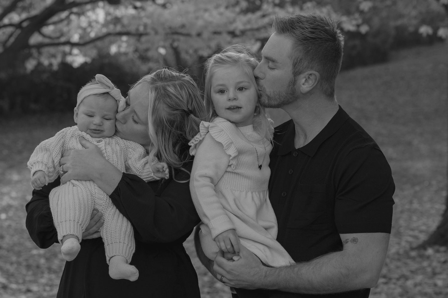 Black and white portrait of parents kissing their two young daughters outdoors in park.