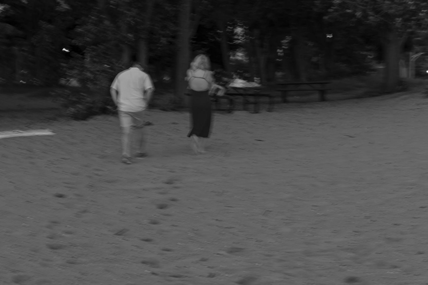 A black and white photo of a couple walking along a sandy beach park at night.