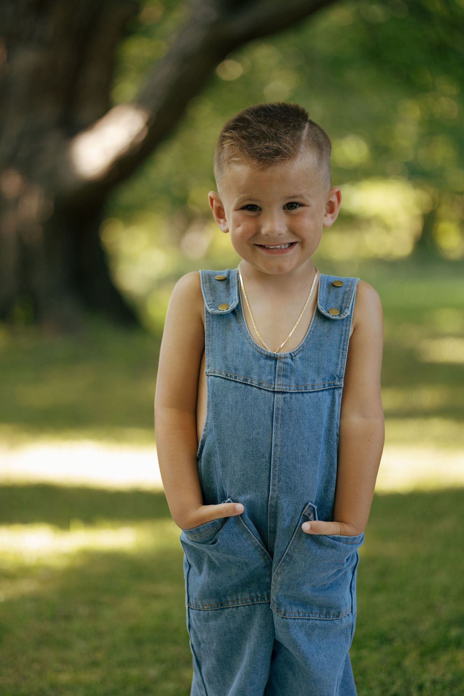 Smiling boy in denim overalls stands with hands in pockets under a large tree in park.