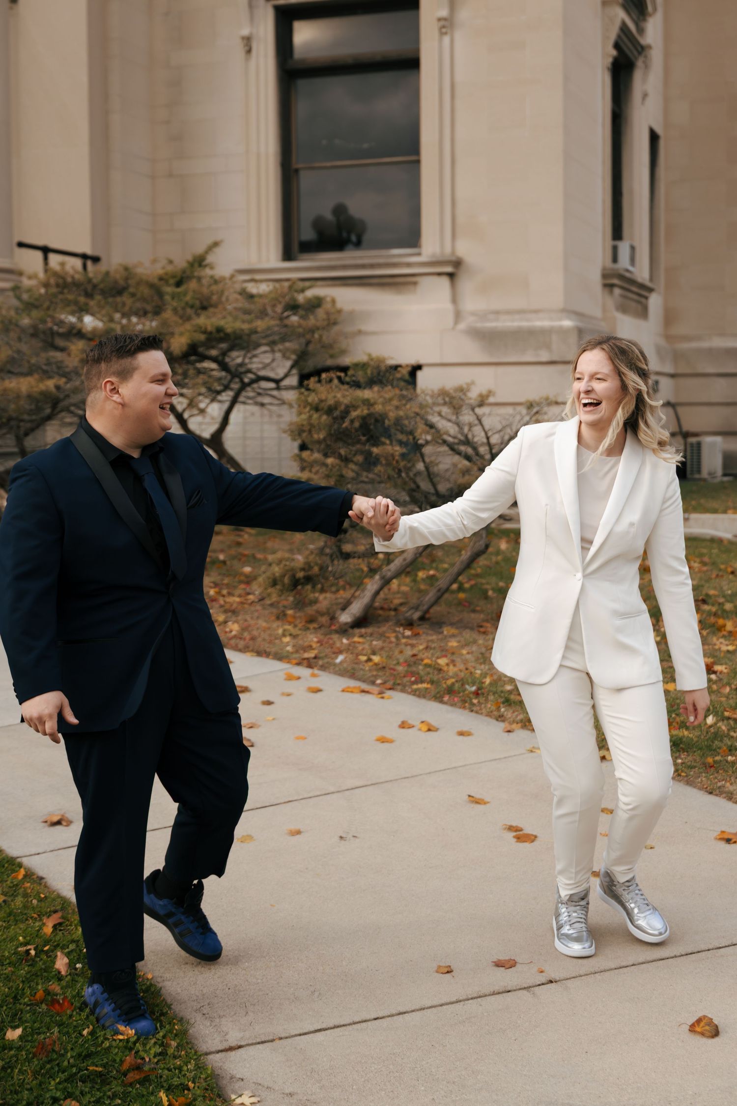 Joyful same-sex couple in black and white suits holding hands laughing outside courthouse.