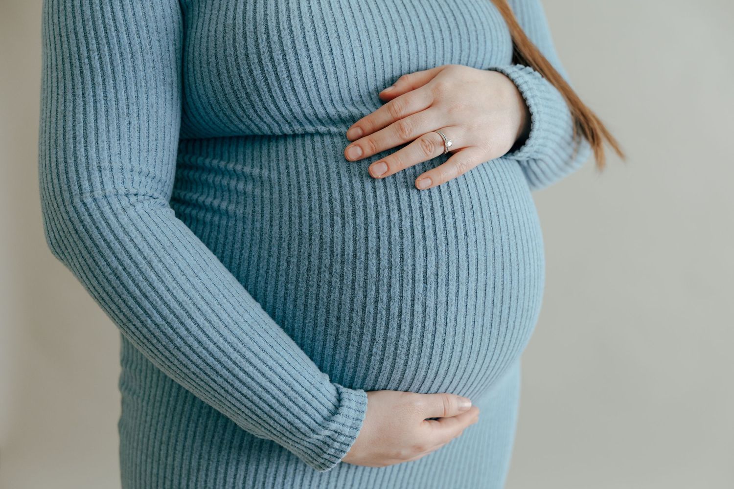 Close-up of pregnant woman in blue ribbed dress cradling baby bump with both hands.