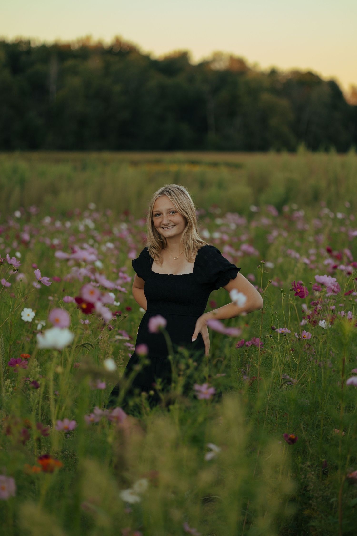 Smiling blonde teen girl posing in vibrant pink cosmos wildflower field at sunset.