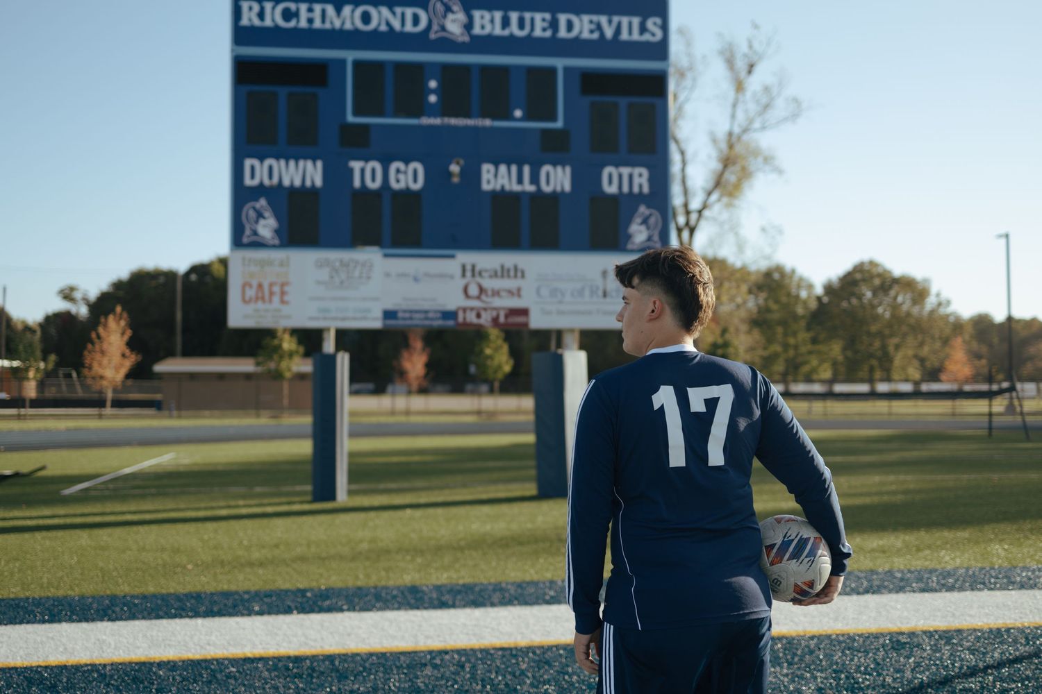 Soccer player #17 stands with ball facing Richmond Blue Devils scoreboard on turf field.