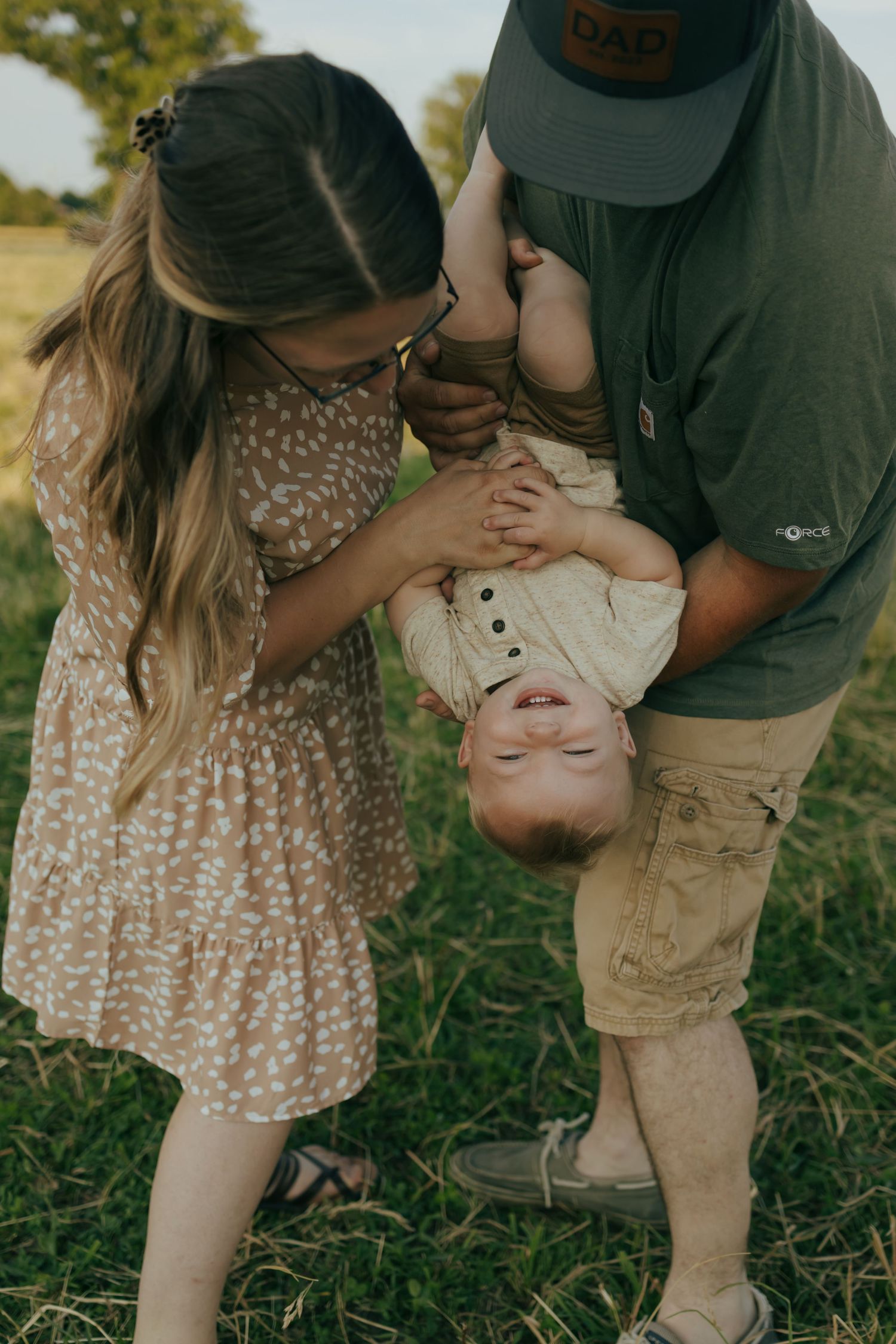 Father holding toddler upside down while sister kisses child in sunny outdoor field.