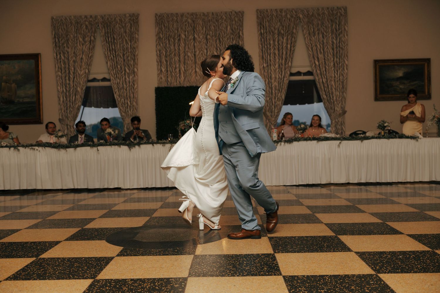 Groom lifting bride during emotional first dance at wedding reception on checkered dance floor.