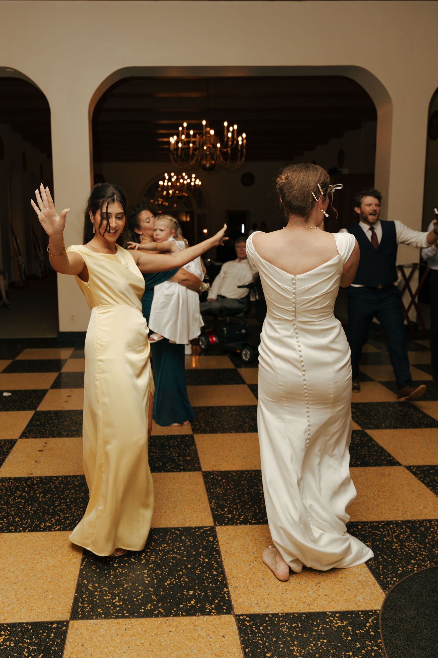 Bride in satin gown dancing with bridesmaid in yellow dress at wedding reception.