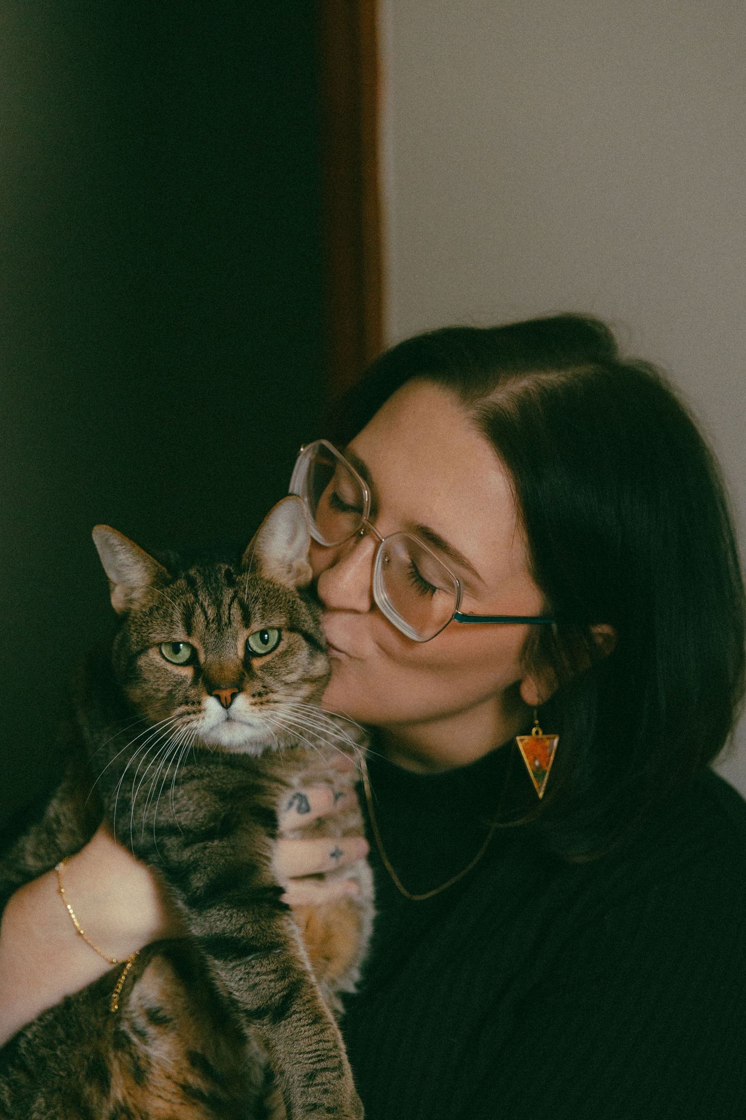 Woman with glasses and triangle earring kissing a tabby cat with green eyes indoors.