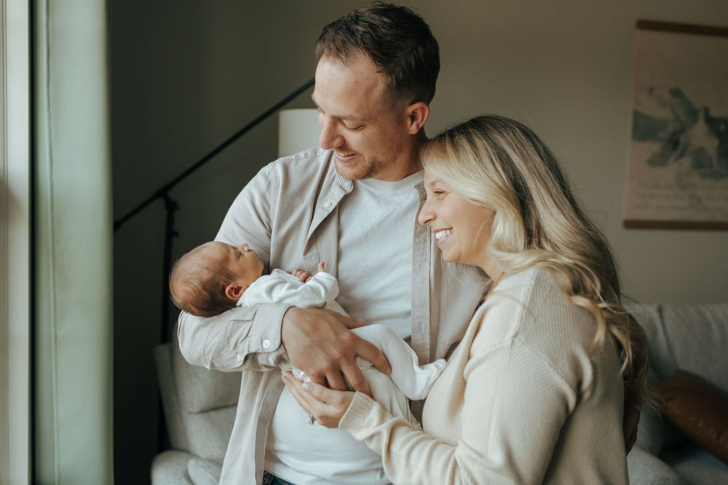 Happy couple smiling and holding newborn baby near window in cozy home setting.