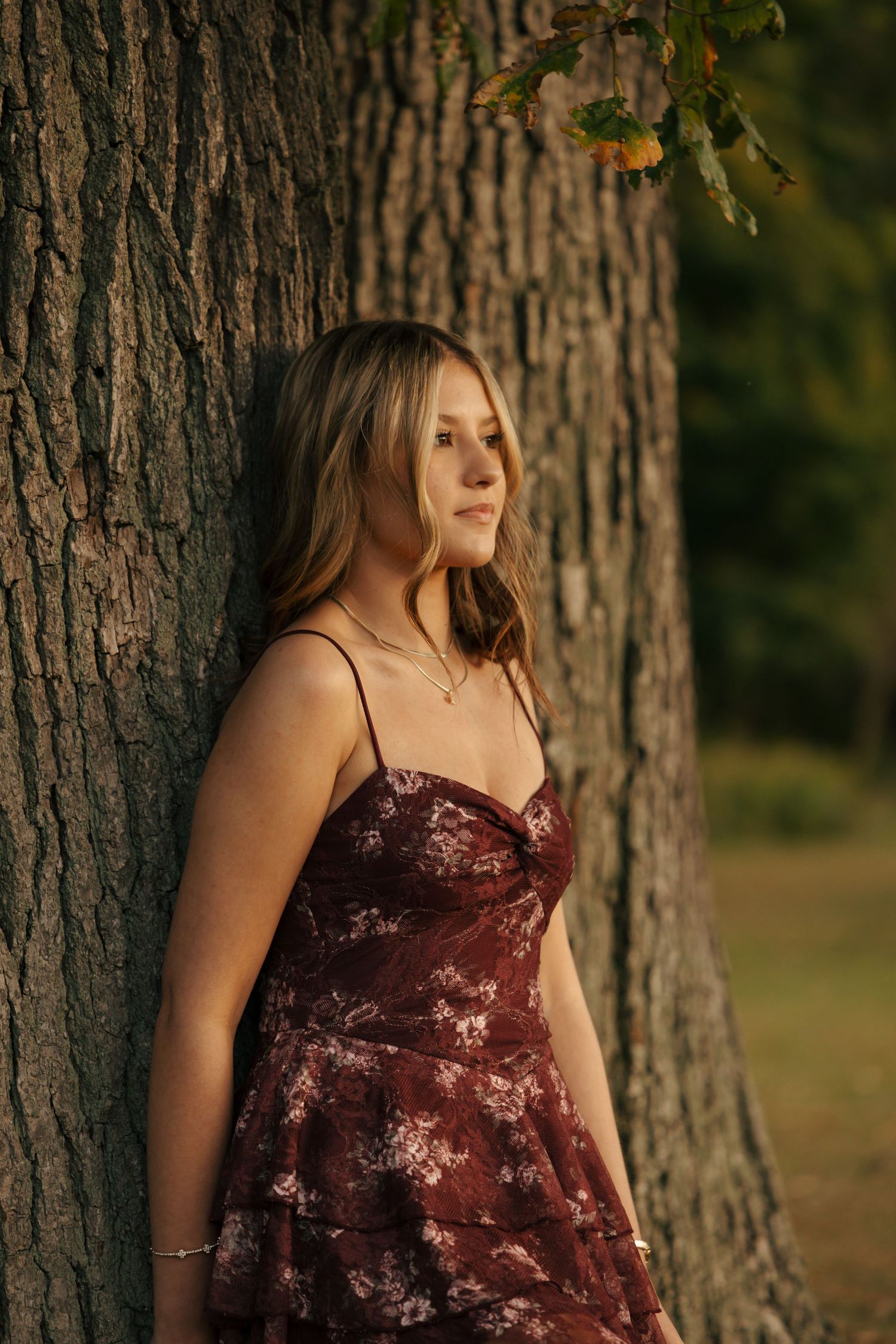 Young woman in burgundy floral dress gazing away while leaning against a large tree.