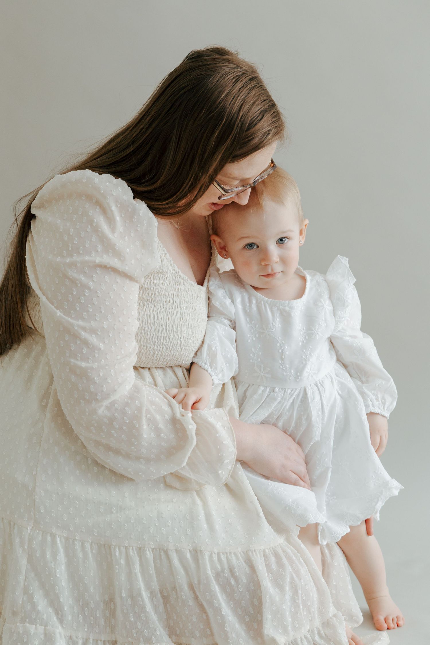 Pregnant woman in white dotted dress tenderly kissing forehead of baby on her lap.