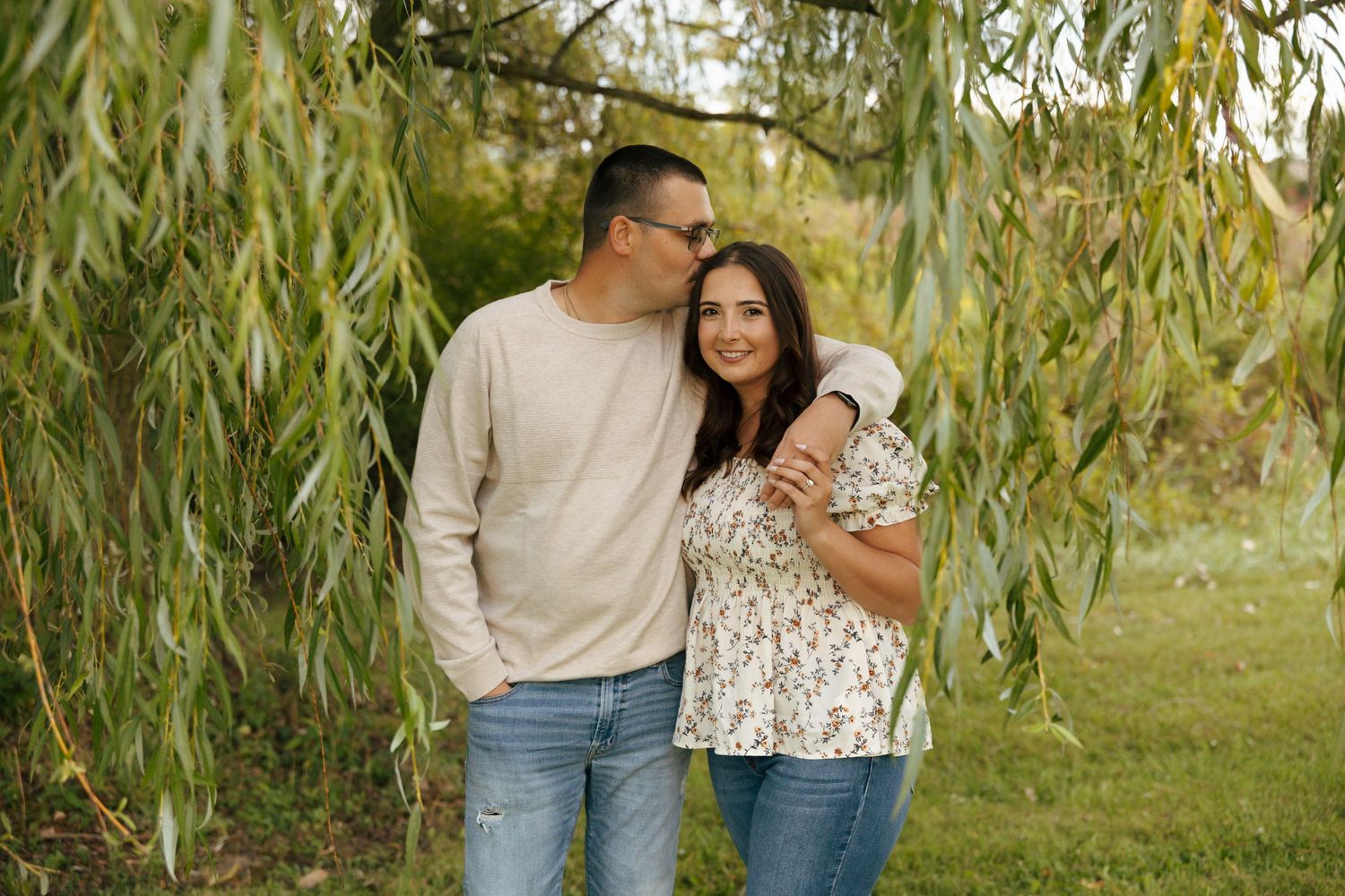 Couple posing together under weeping willow branches, man kissing womans cheek outdoors.