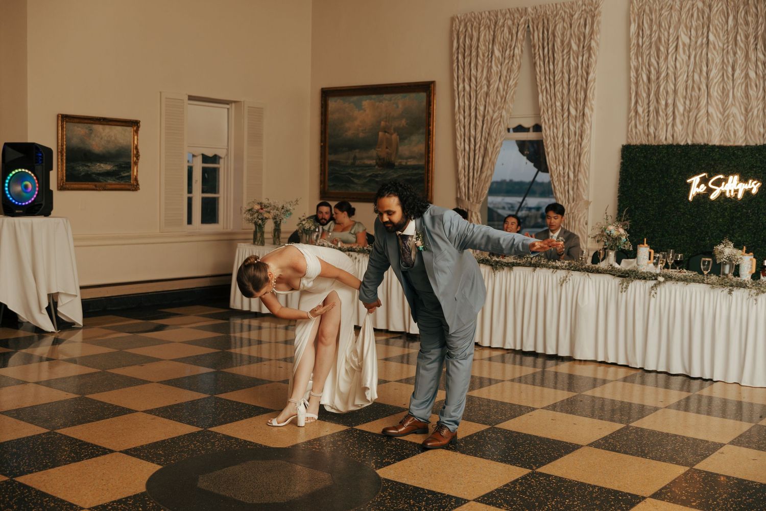 Groom bending bride in dramatic dance dip at wedding reception on checkered ballroom floor.