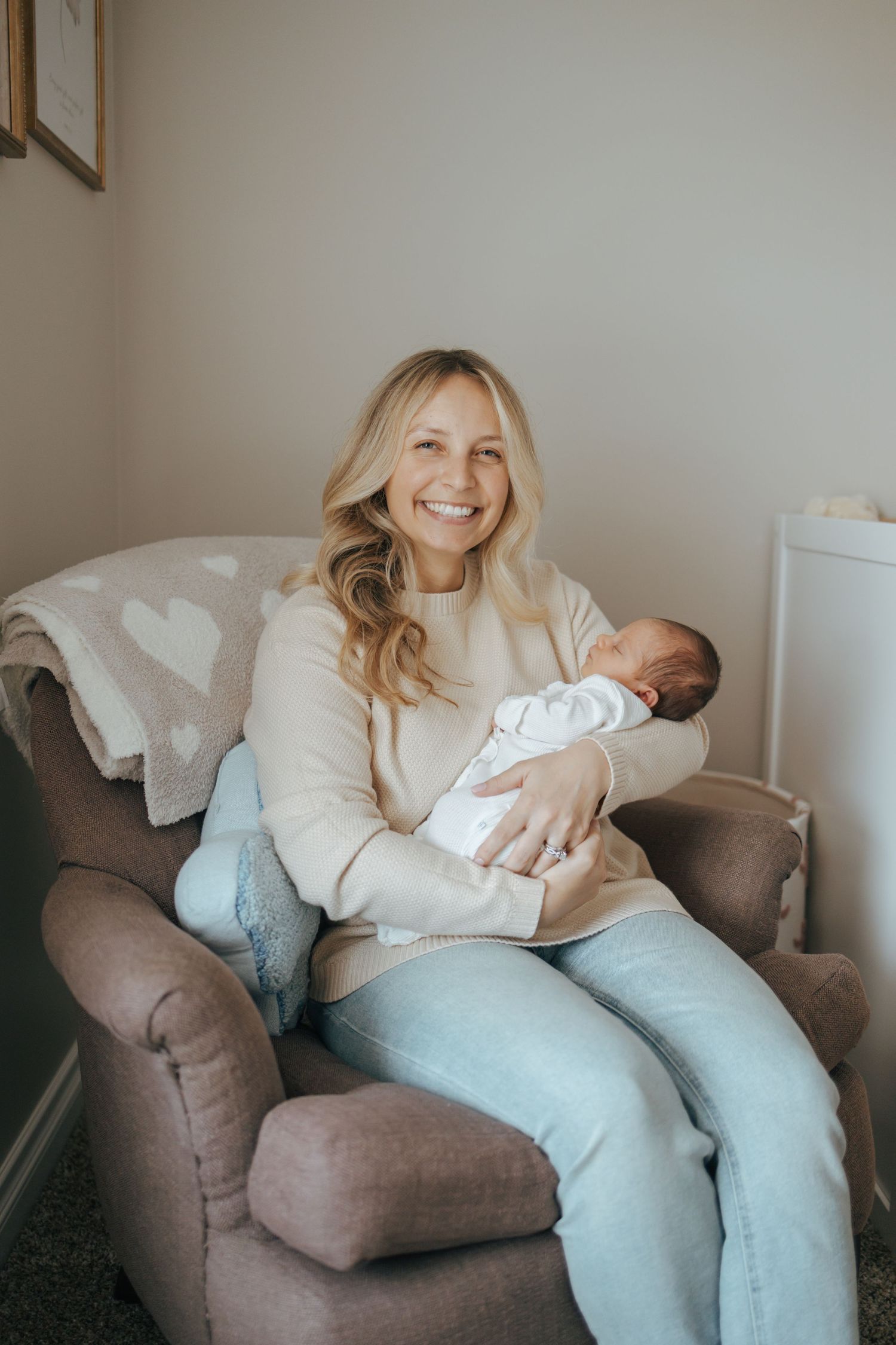 Smiling blonde mother sitting in armchair holding sleeping newborn baby in cream nursery room.