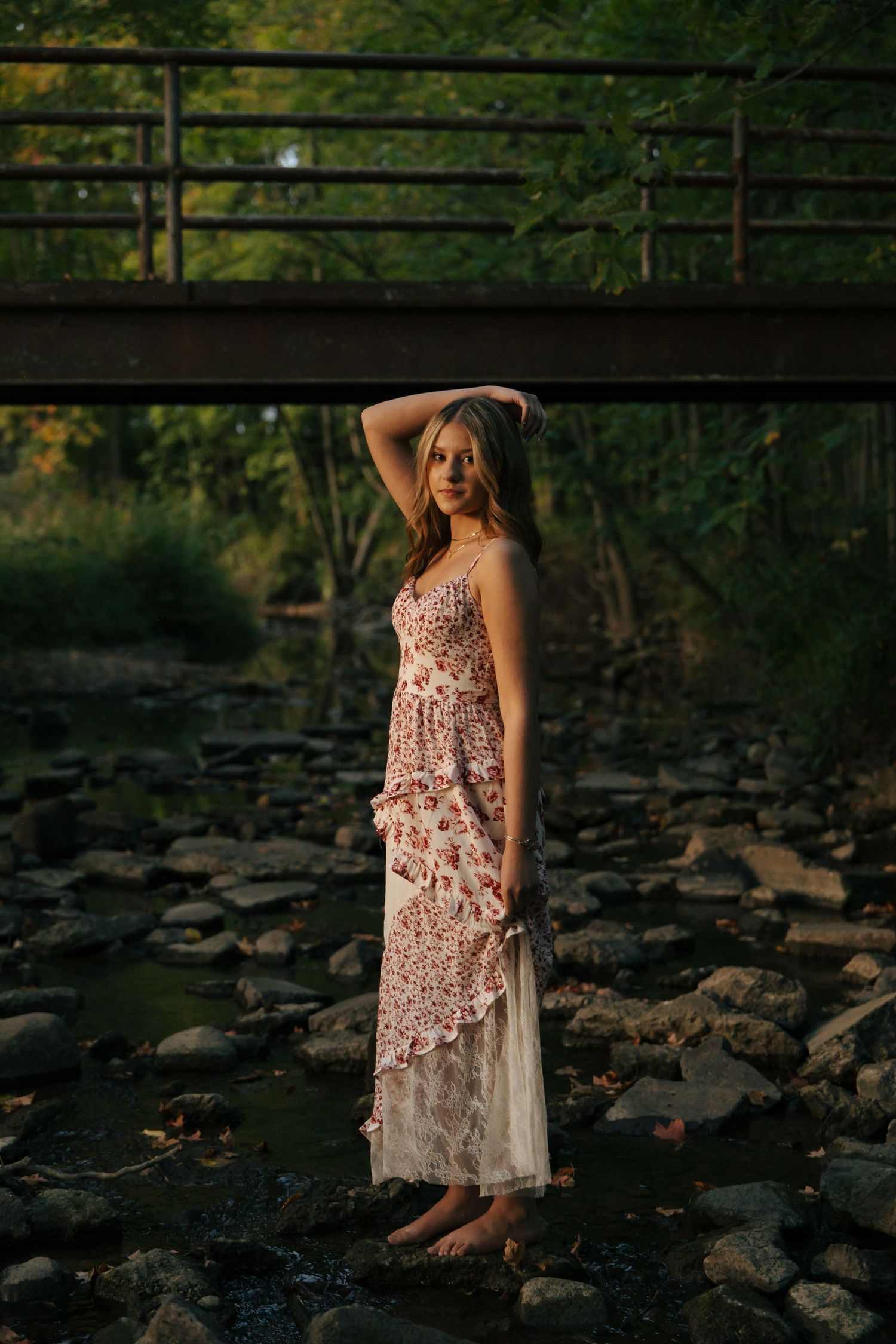 Young woman in floral maxi dress standing barefoot on rocks beneath a bridge at sunset.