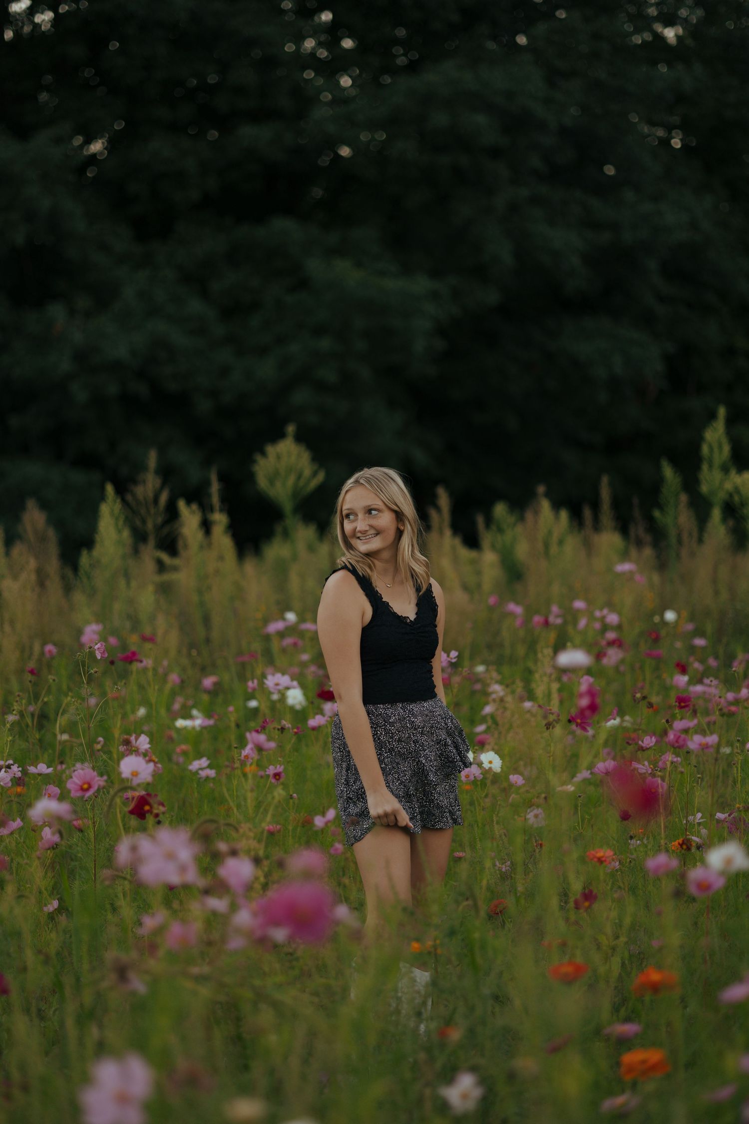 Smiling blonde teen girl in black tank top and floral skirt standing in wildflower field.