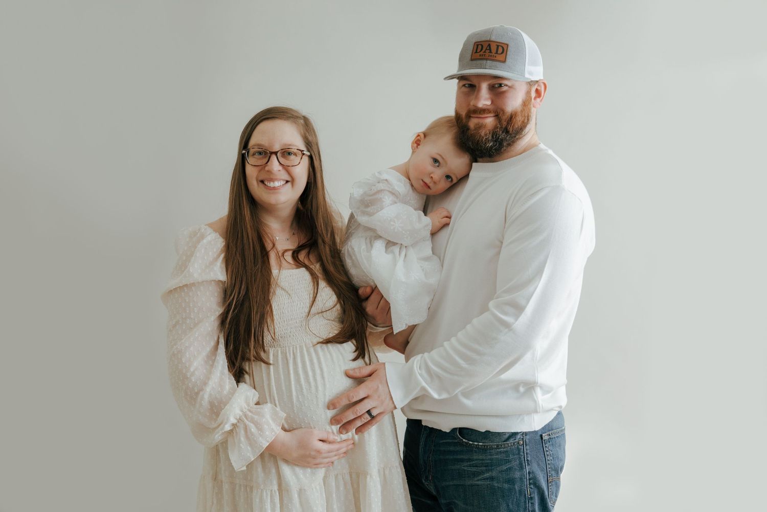 Smiling pregnant woman and bearded man holding toddler in white outfit for maternity photo.