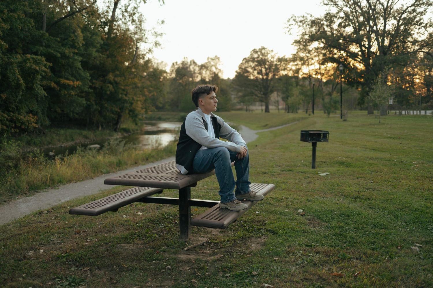 Young man sits alone on park picnic table bench at golden hour near a pond.