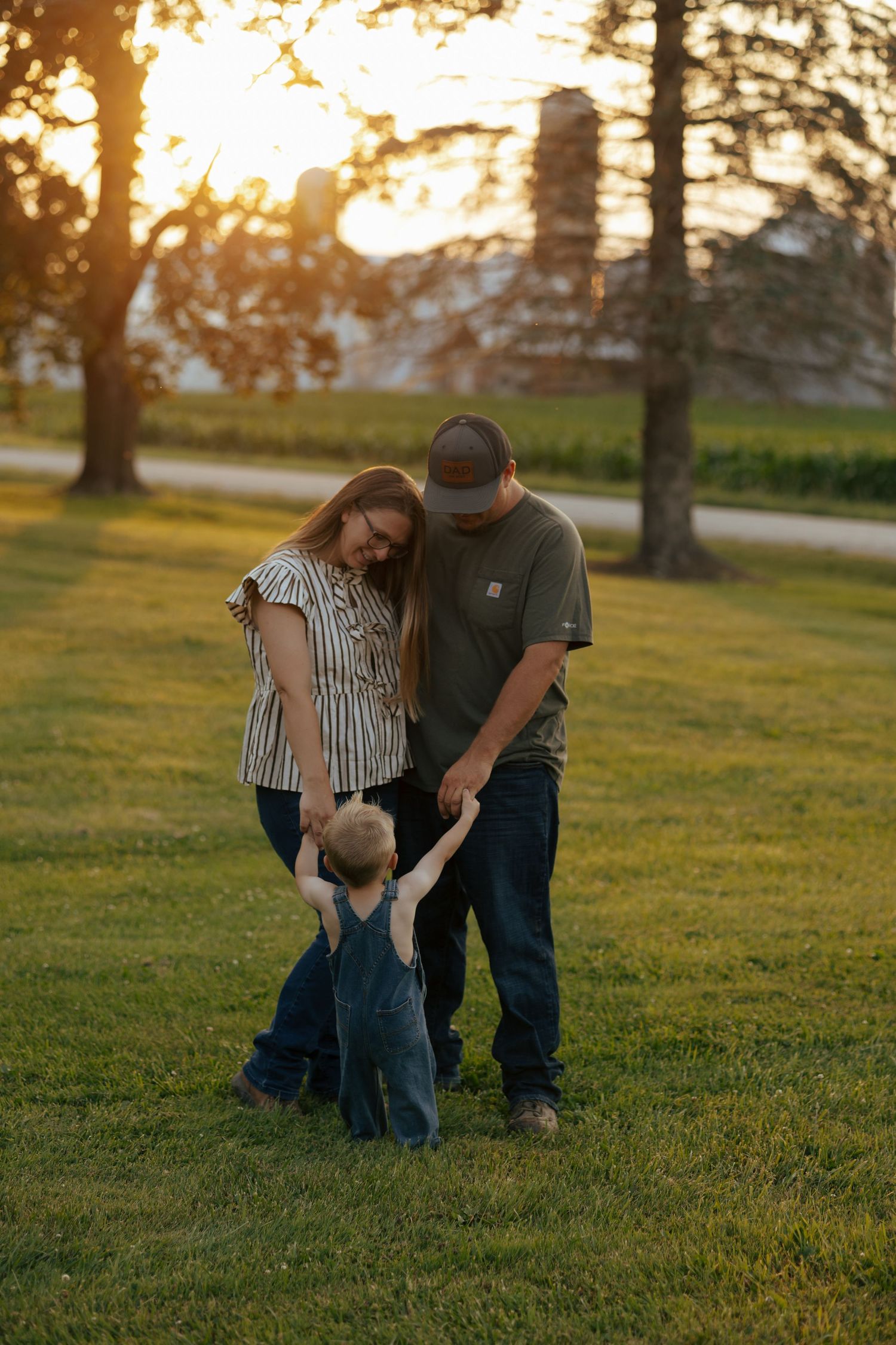 Family of three at golden hour, parents holding toddlers hands while walking on lush green grass.