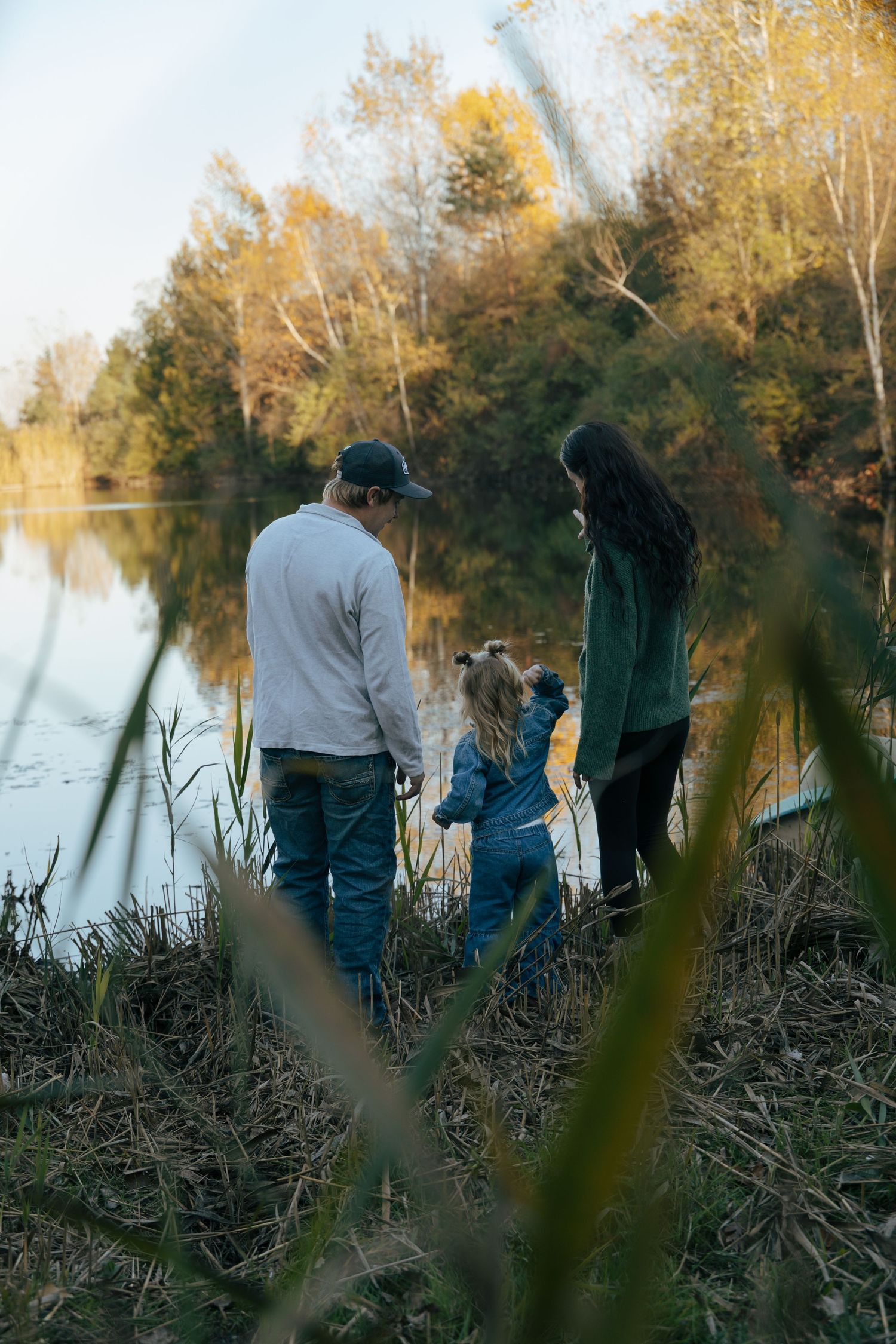 Family of three holding hands while standing at the edge of a calm reflective lake in autumn.