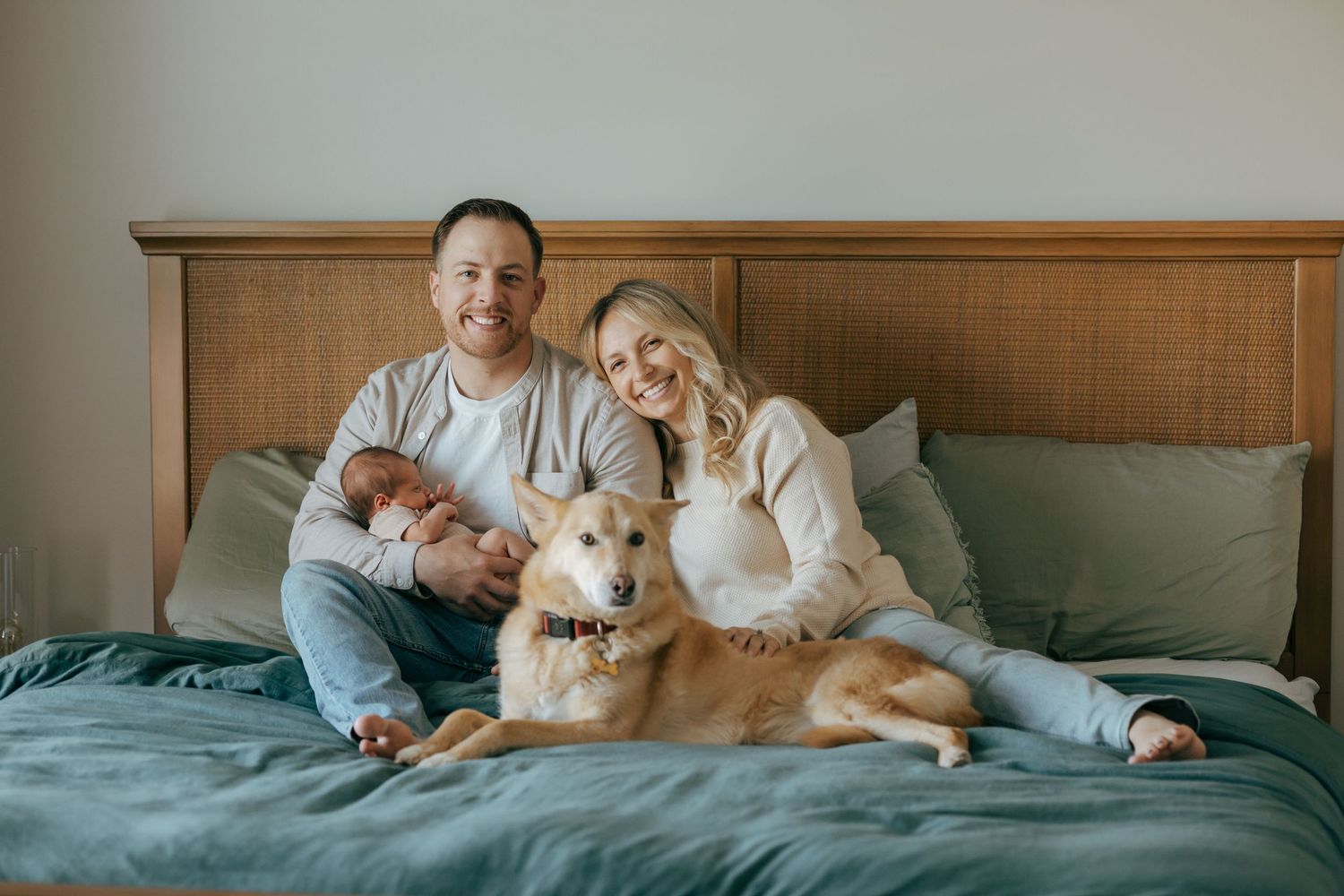 Smiling couple sitting on a green bed with a newborn baby and golden dog.