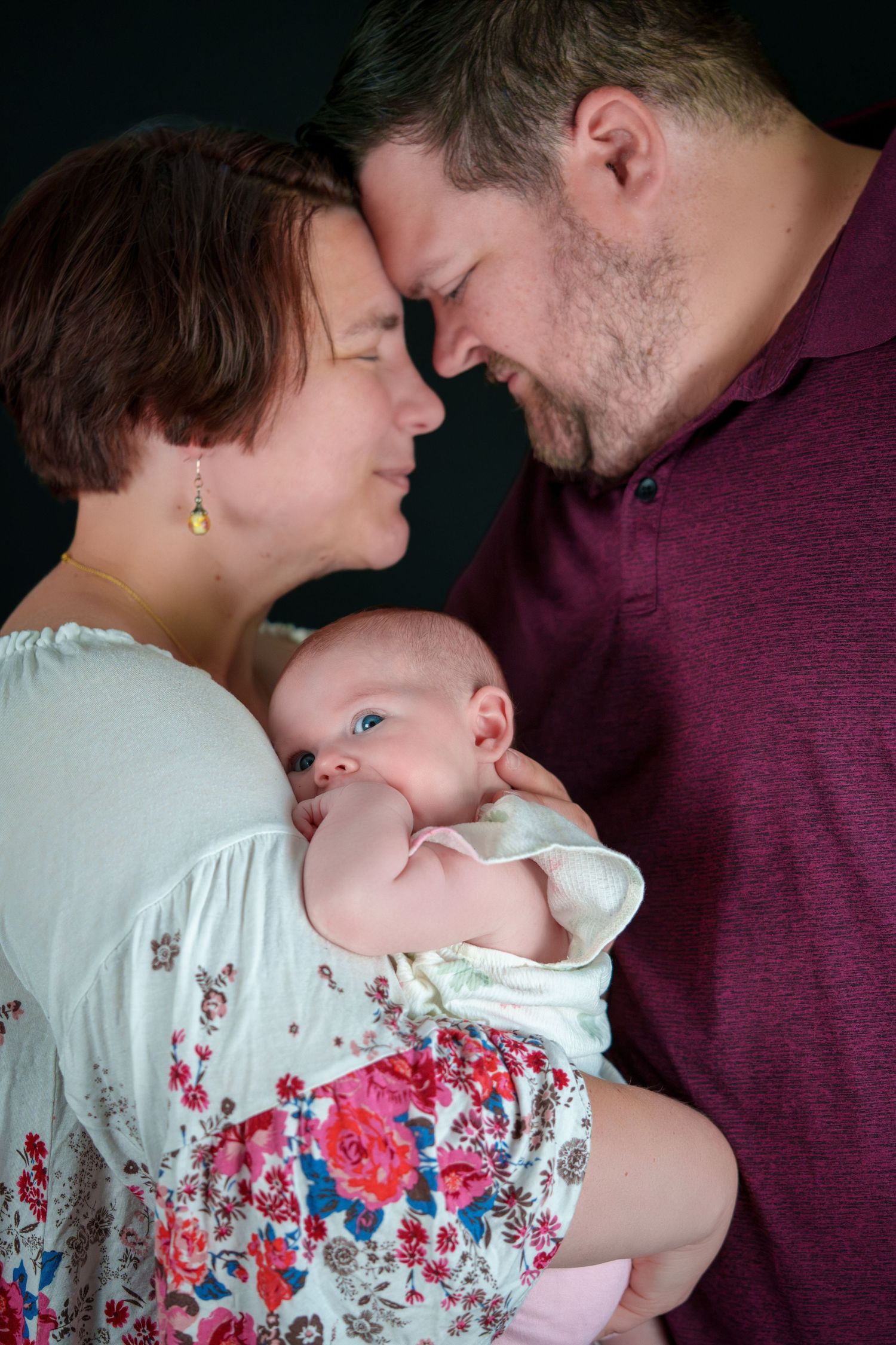 A couple with foreheads touching lovingly hold their newborn baby, smiling in a studio portrait.