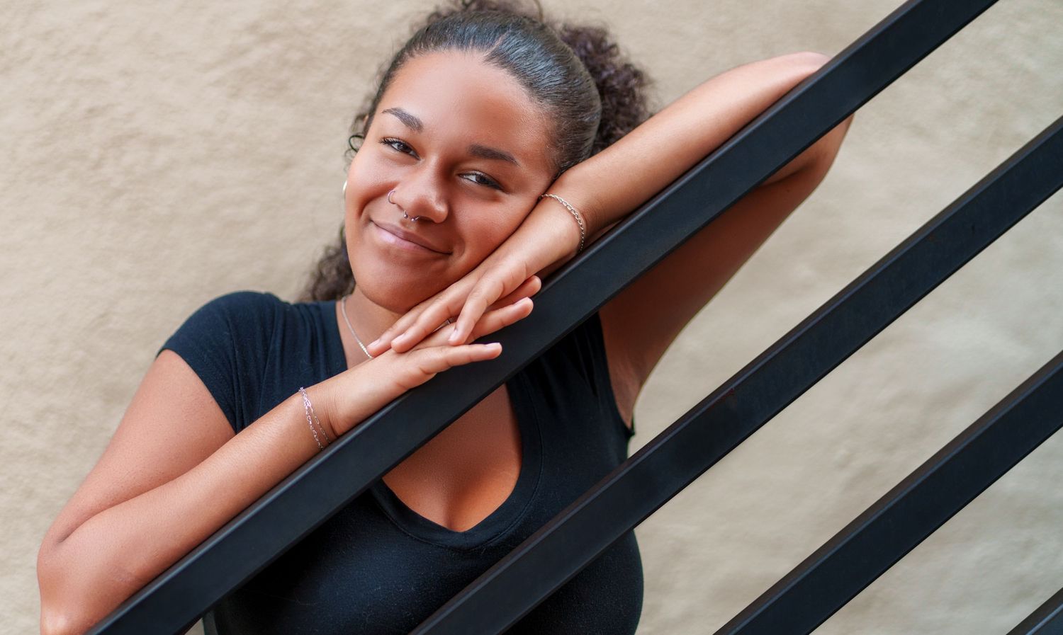 Young woman with curly hair smiling and leaning on black metal railing outdoors.