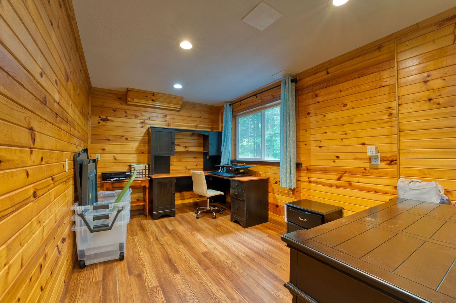 Cozy wood-paneled home office with hardwood floors, black desk, office chair, and natural light from a window.