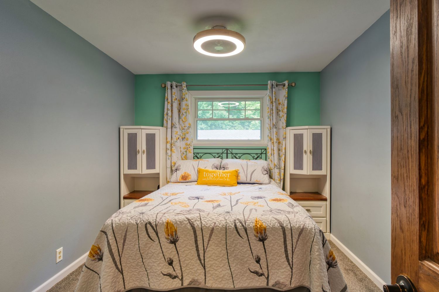 Cozy bedroom with blue-green walls, floral bedspread, yellow pillows, twin nightstands, and a window with patterned curtains.