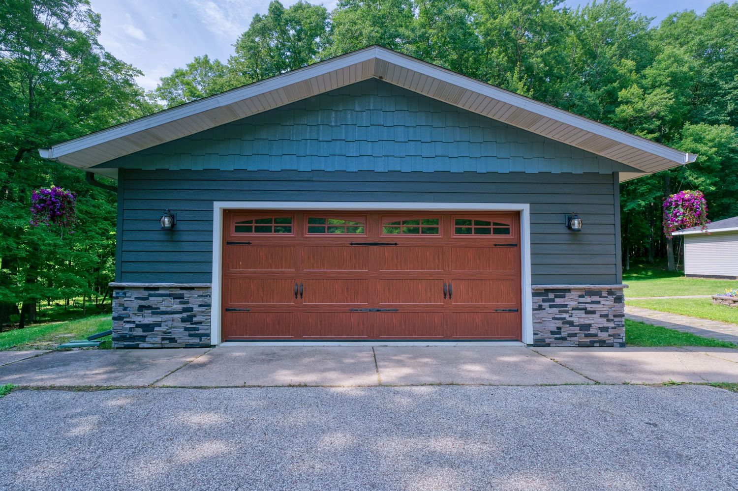 Blue garage with brown wooden door, stone accents, hanging flower baskets, and lush green trees in background.