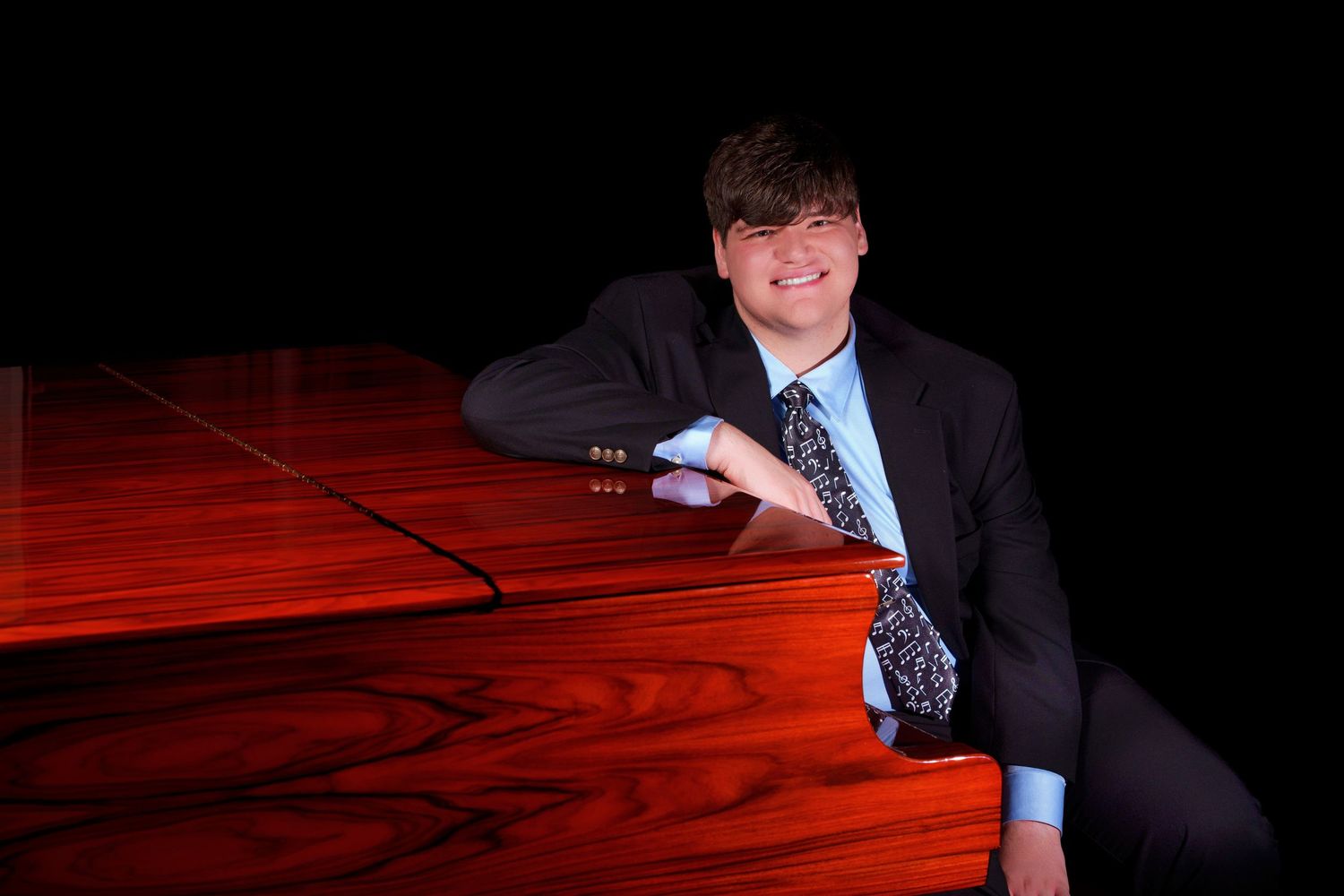 Young man in suit and tie posing confidently beside a polished red grand piano against dark background.