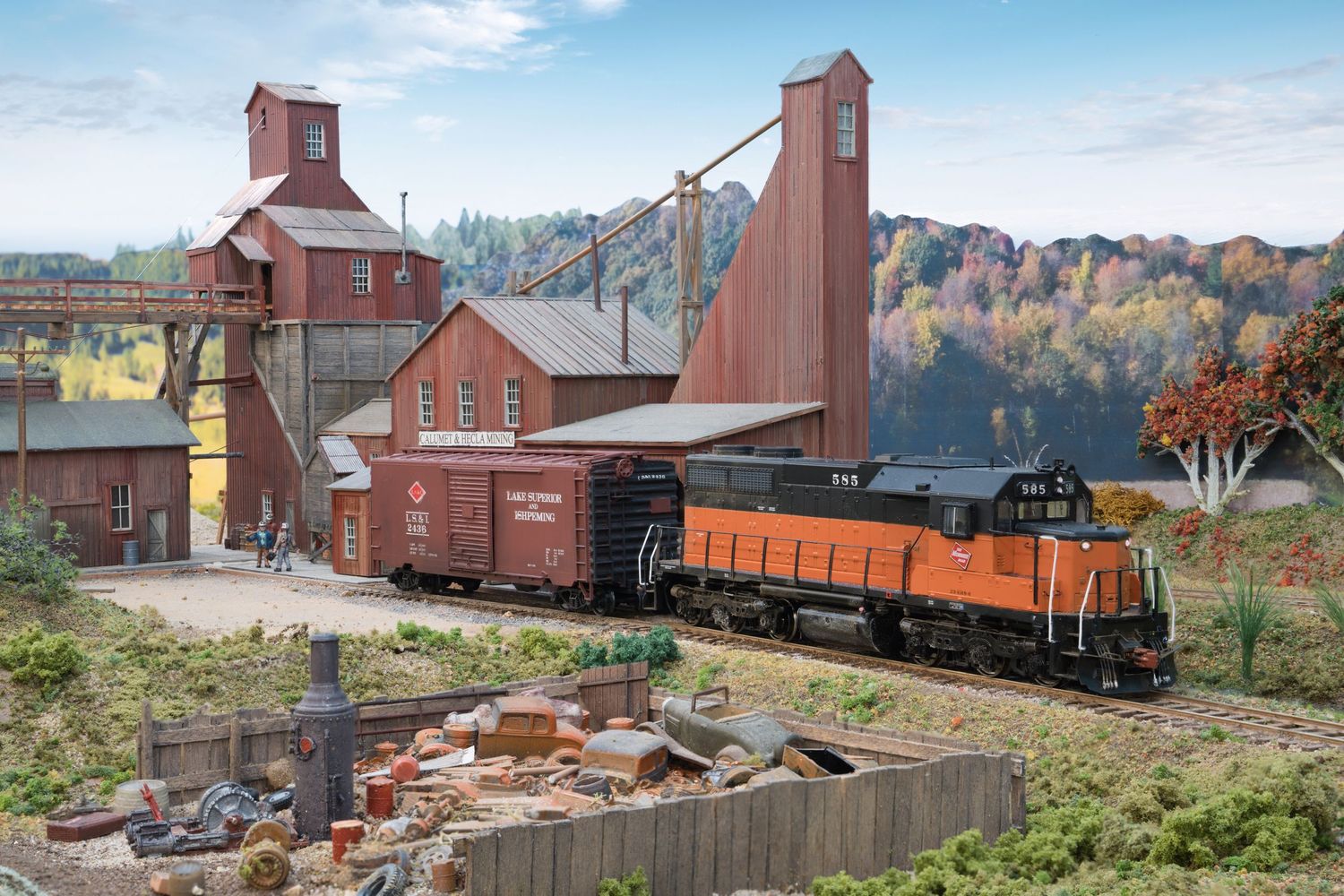 A series of vintage train cars and industrial buildings line up along railroad tracks in a scenic autumn landscape.