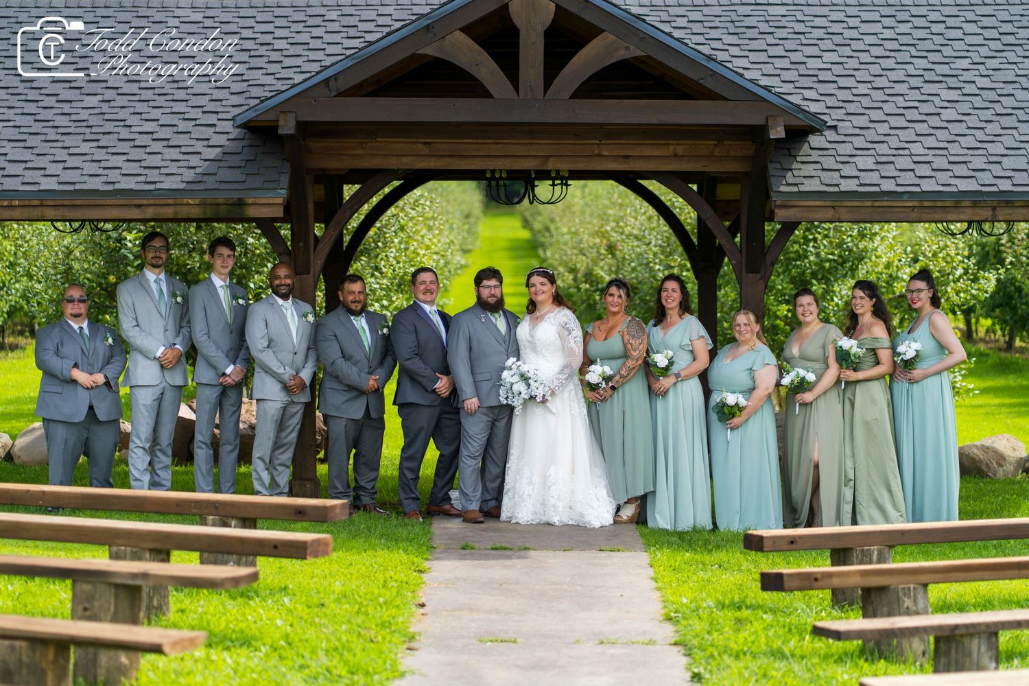 A wedding party in gray and white attire poses together outdoors in front of wooden benches and green scenery.