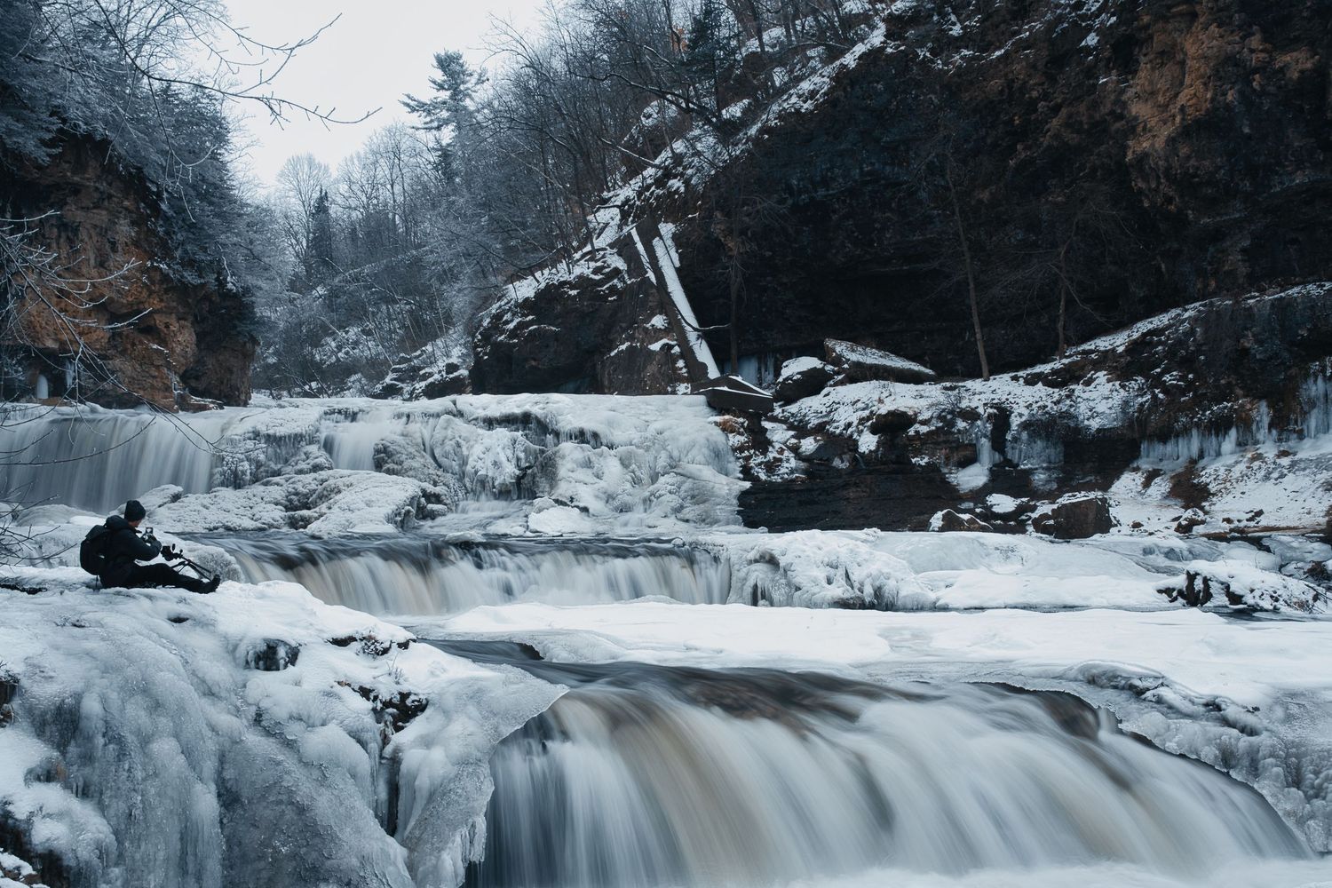 A frozen waterfall cascades down snow-covered rocks in a winter landscape.