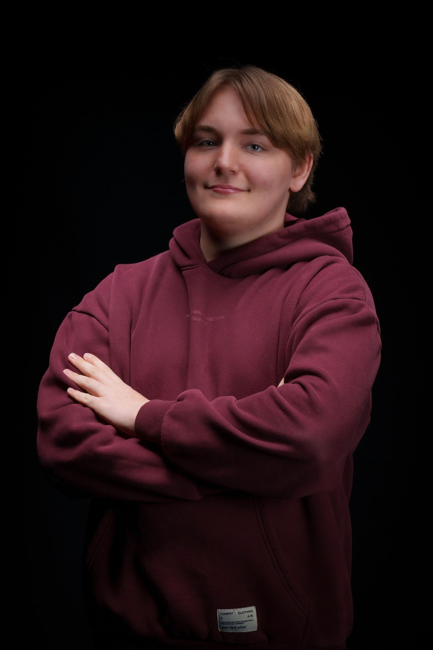 Young person in maroon hoodie with arms crossed smiling against black background.