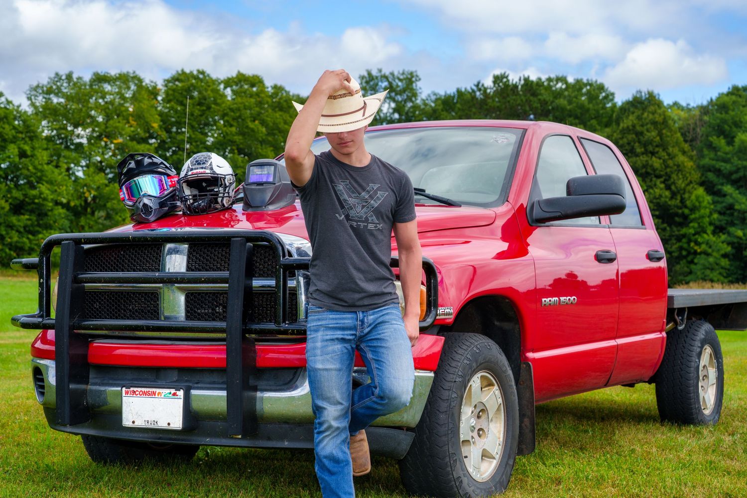 Man in cowboy hat leaning against red pickup truck on green field with racing helmets in truck bed.