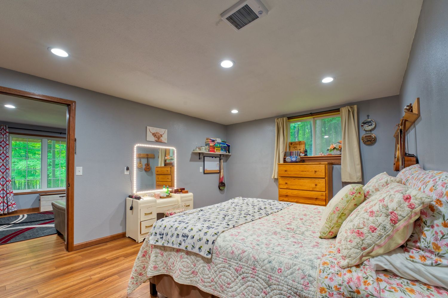 Cozy bedroom with hardwood floors, recessed lighting, floral bedding, wooden dresser, and windows with green outdoor views.