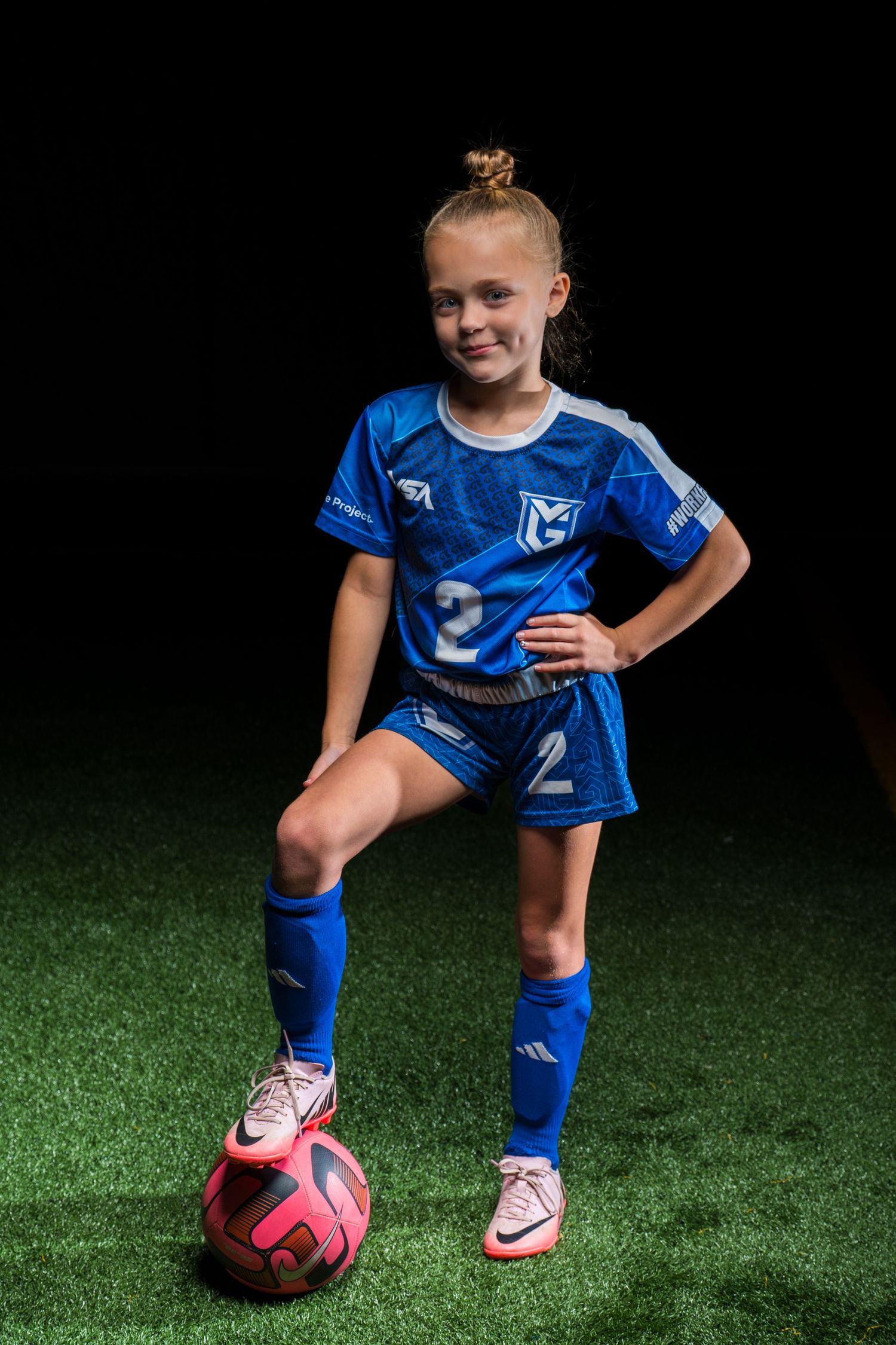 Young soccer player in blue uniform poses with pink ball on artificial turf in dramatic lighting.