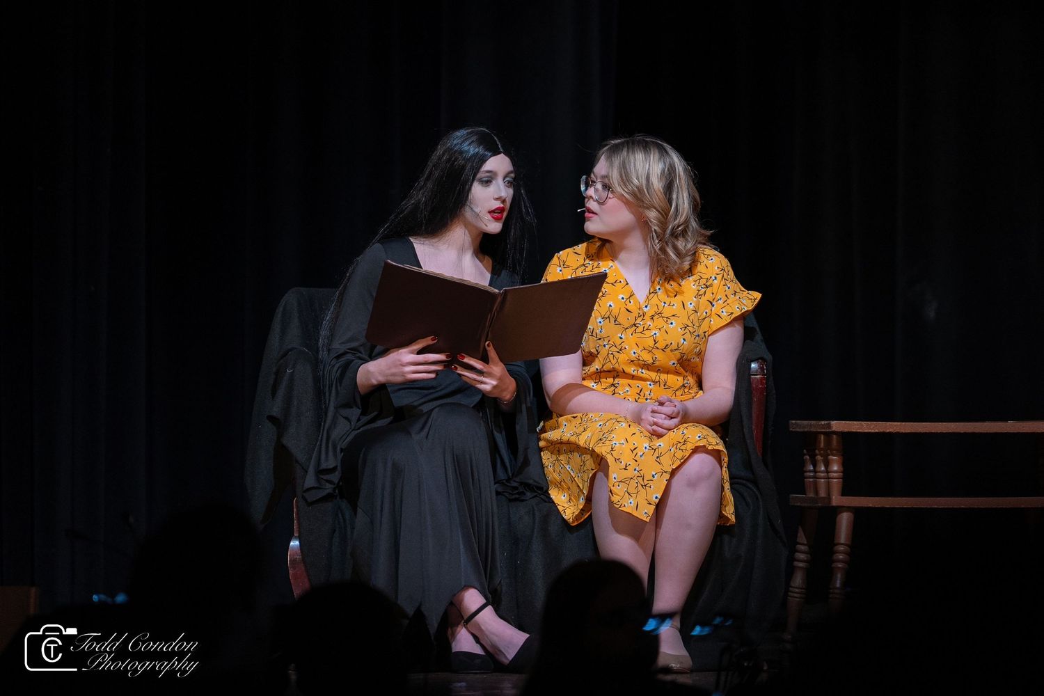 Theater actors performing on a dimly lit stage with one wearing a yellow dress and another in black.
