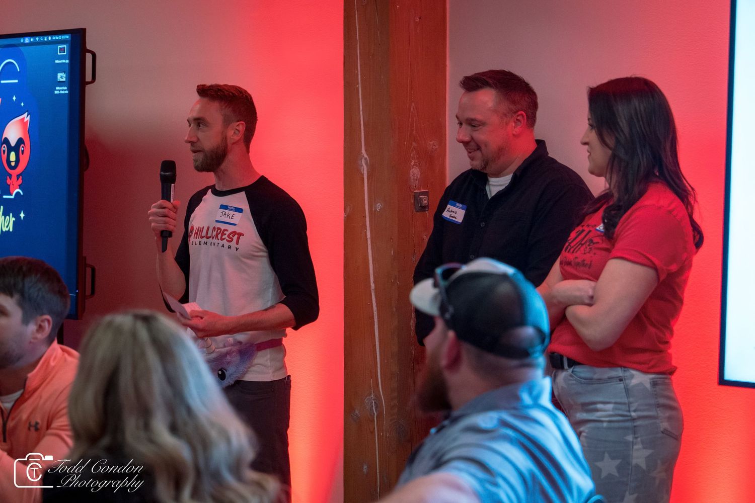 Three people presenting on stage in a red-lit room, one holding a microphone, at an indoor event.