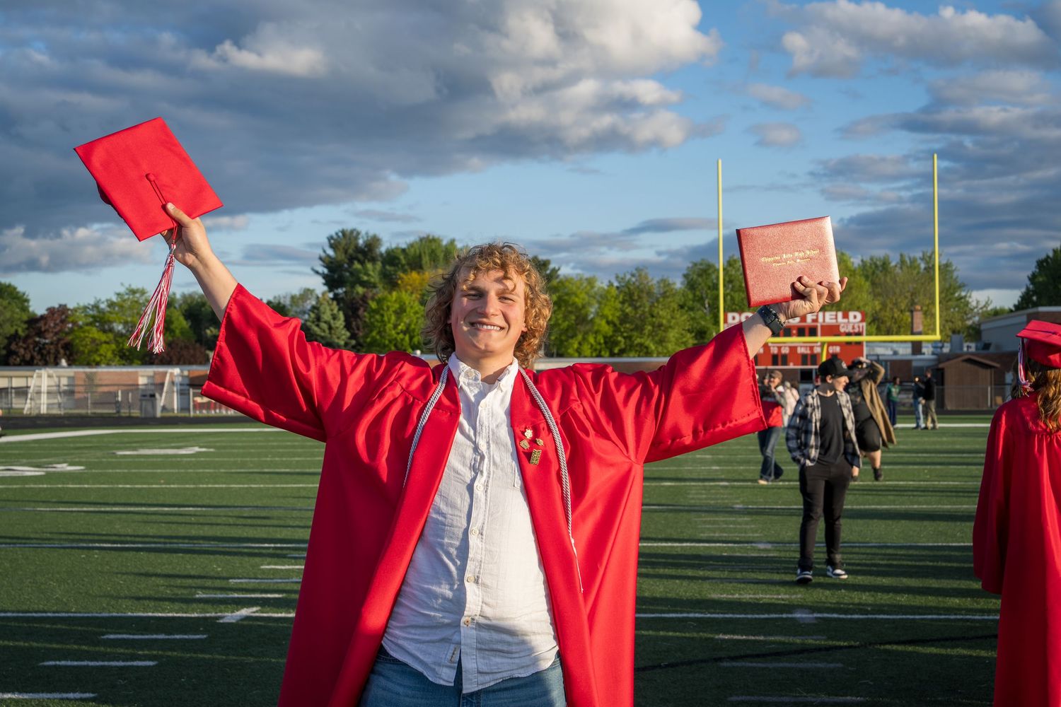 Joyful graduate in red cap and gown celebrates on football field holding diploma at outdoor graduation ceremony.