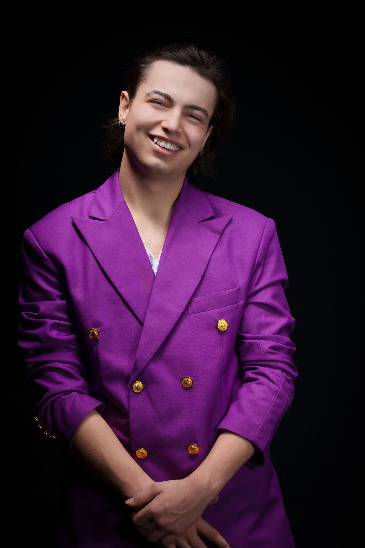 Smiling young man in a bold purple double-breasted blazer with gold buttons, posing against a black background.