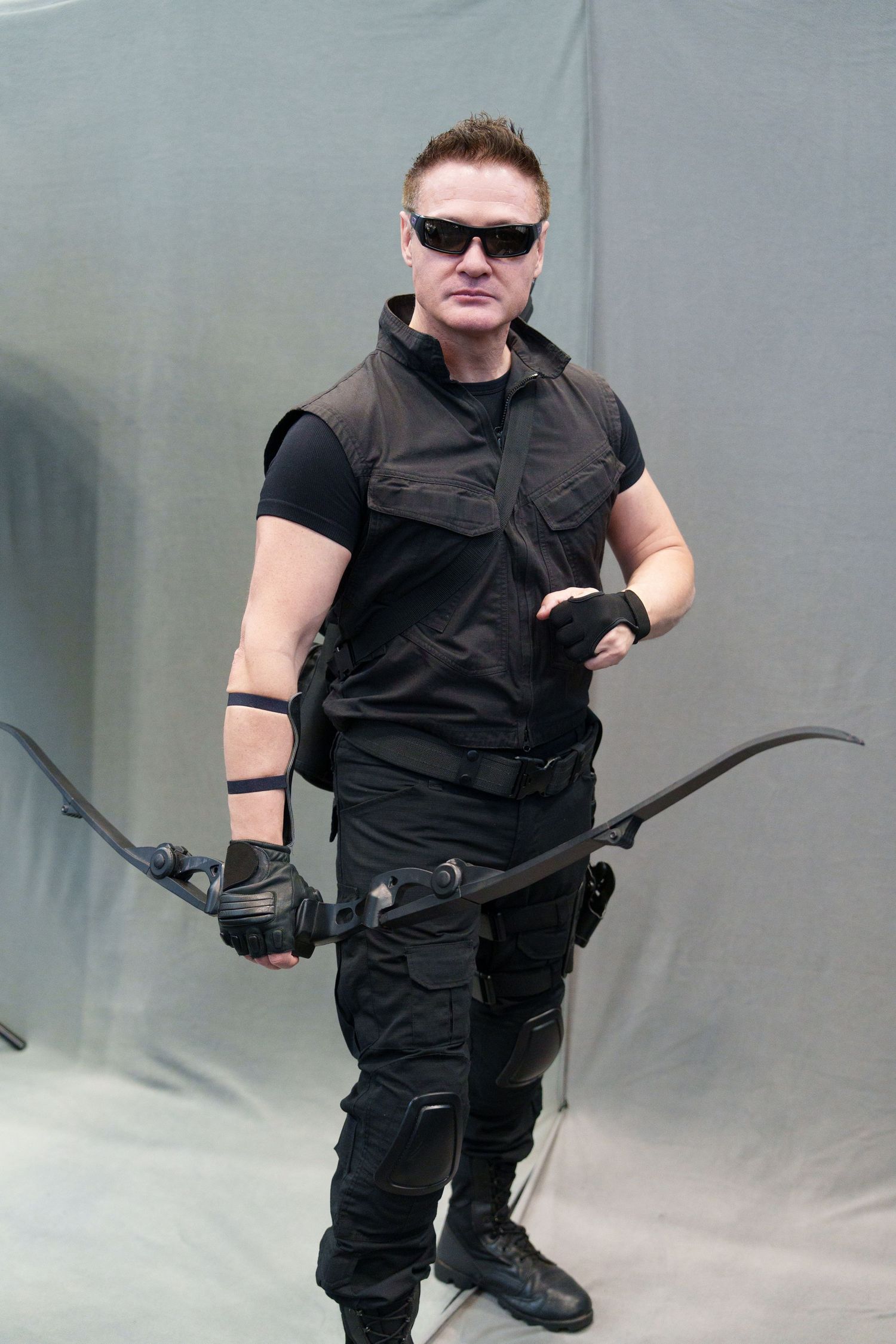 Man in black tactical gear holding a bow, wearing sunglasses, posing as Hawkeye cosplay against gray backdrop.