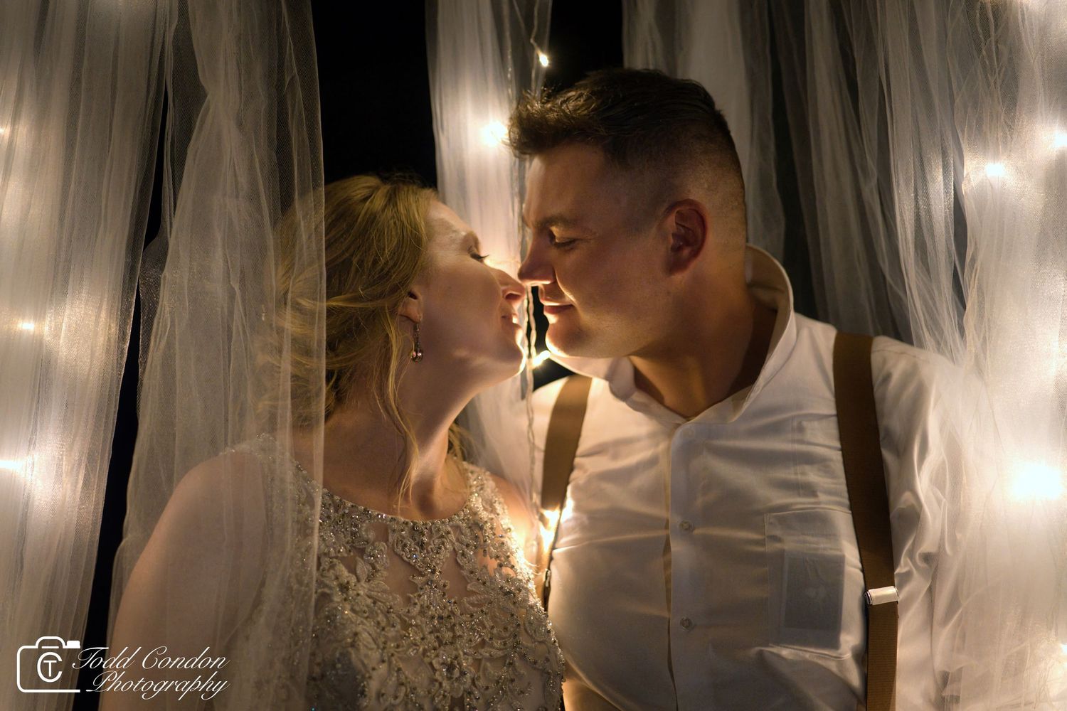A romantic wedding couple shares an intimate moment under soft glowing lights and white fabric drapes.