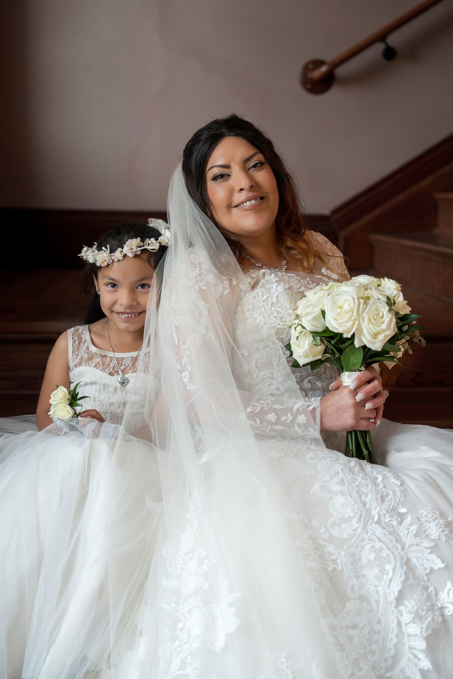 A bride in a white lace wedding dress sits on stairs holding a bouquet of white roses.